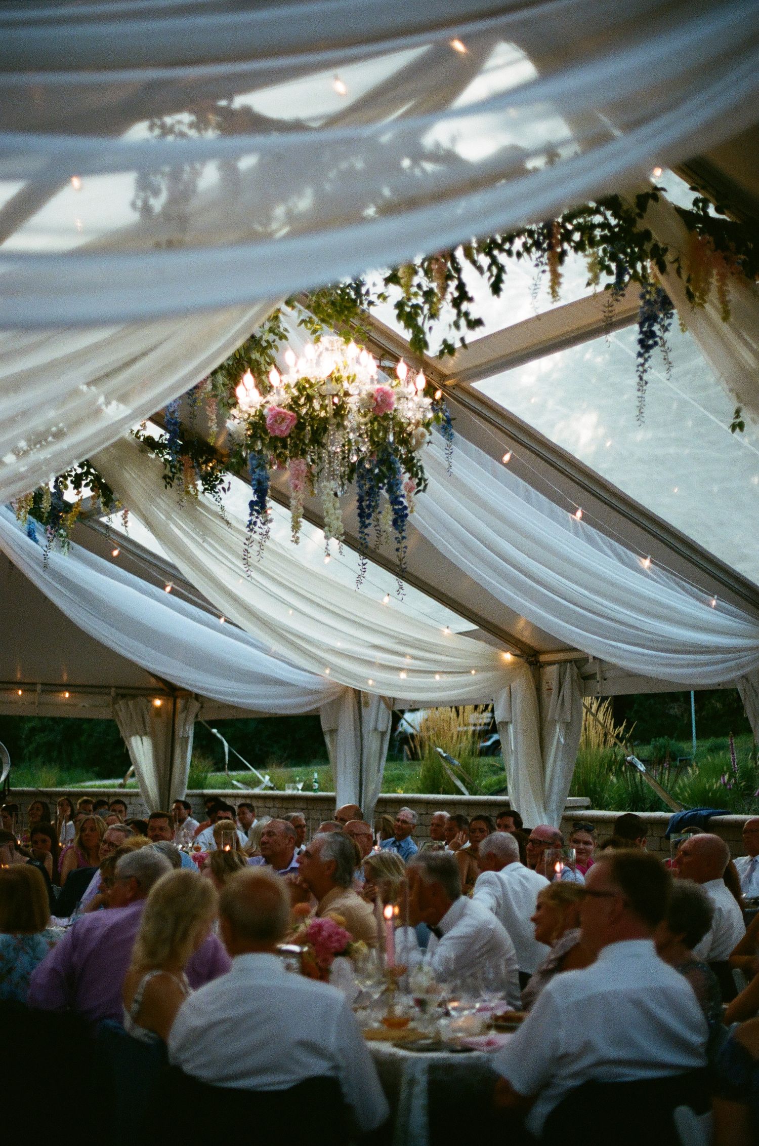 Elegant outdoor wedding reception under a clear tent with white draping, floral arrangements, and guests dining by candlelight.