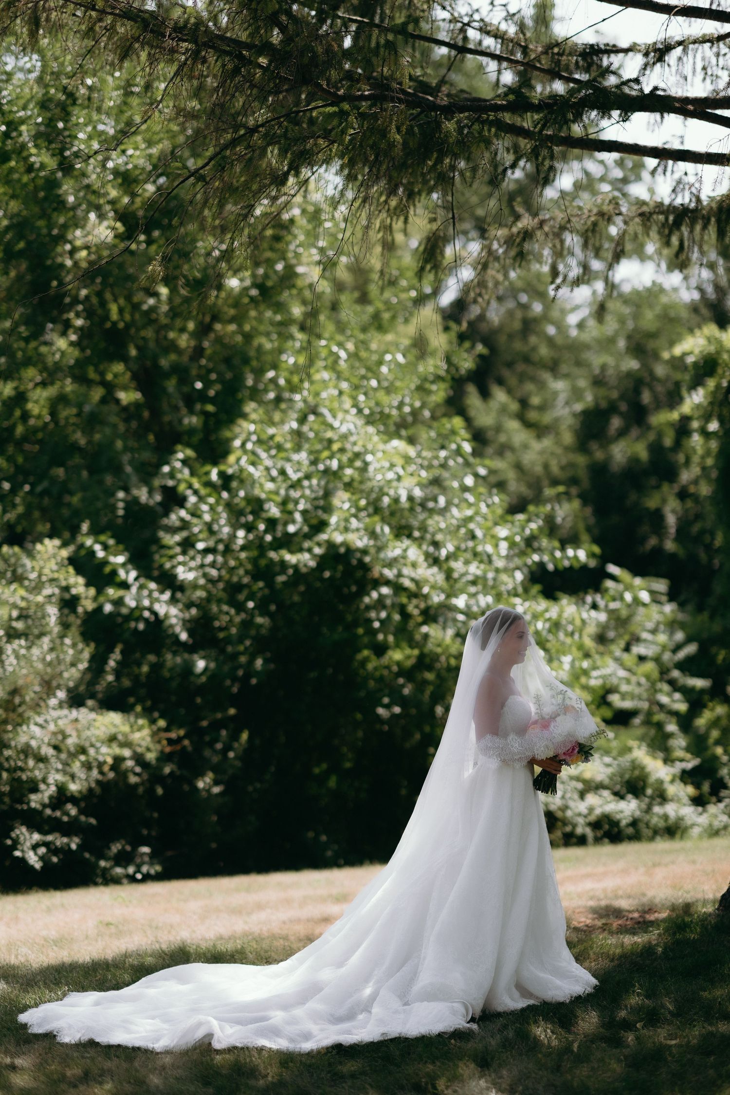 Bride in white gown and veil holding bouquet stands in lush green garden on wedding day.