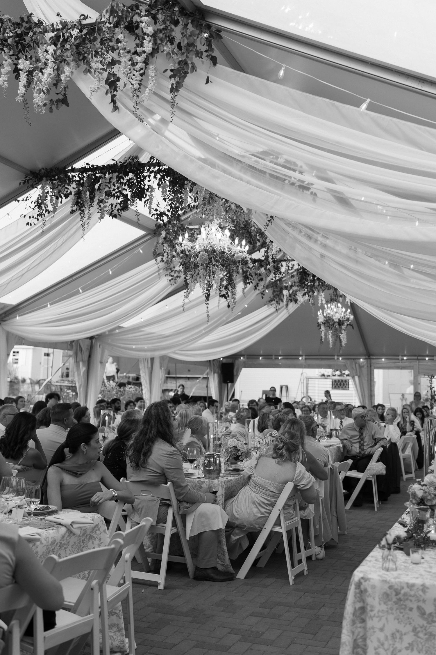 Black and white aerial view of elegant wedding reception under draped tent with chandeliers and seated guests.