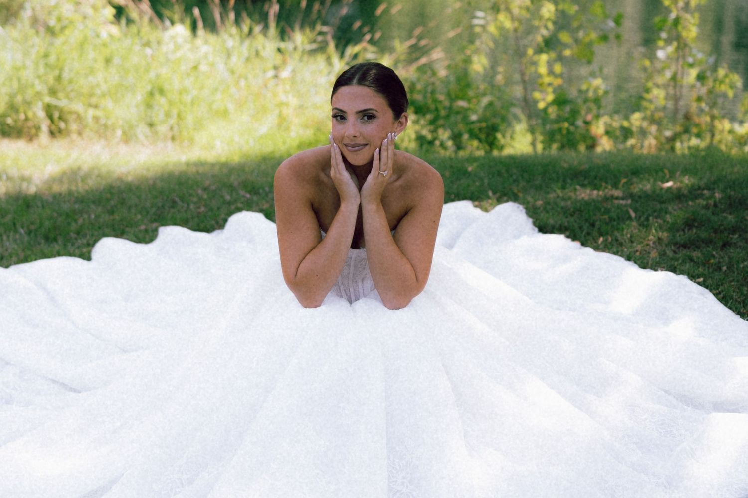 A radiant bride in a voluminous white ball gown poses on green grass surrounded by lush nature.