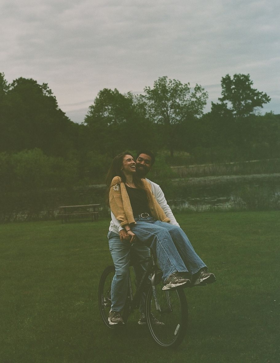 Couple laughing together on a bicycle in a lush green park on a cloudy day.