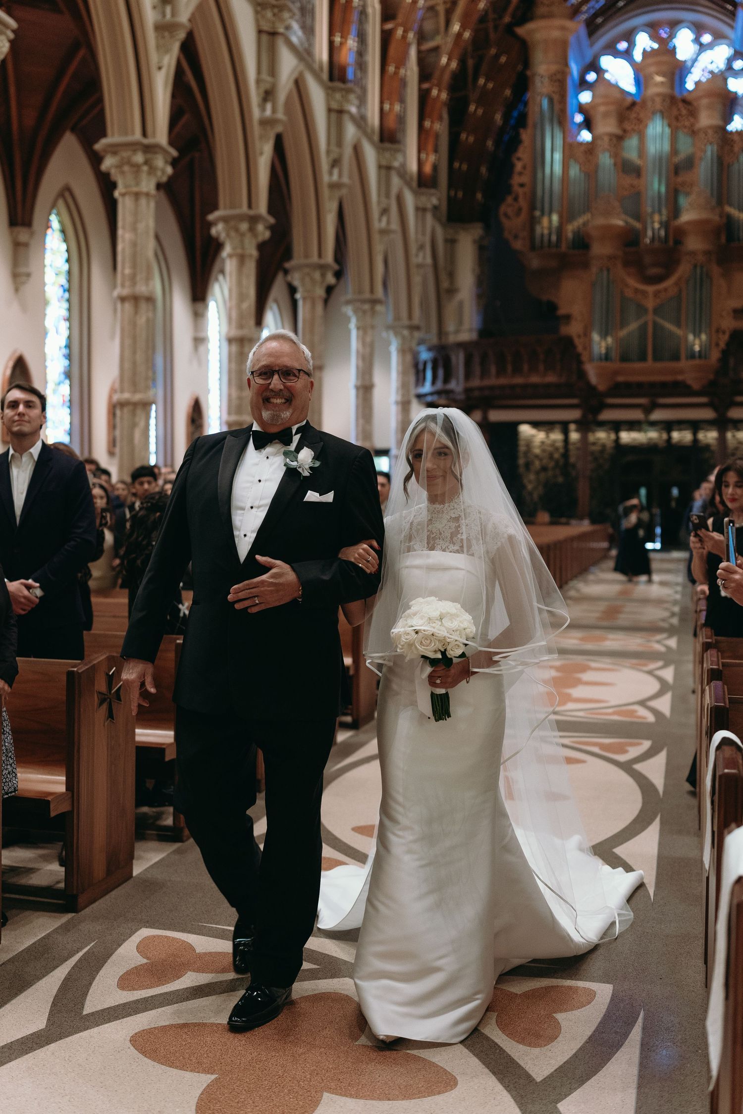 A wedding couple walks down a church aisle with ornate marble floors and gothic architectural details.