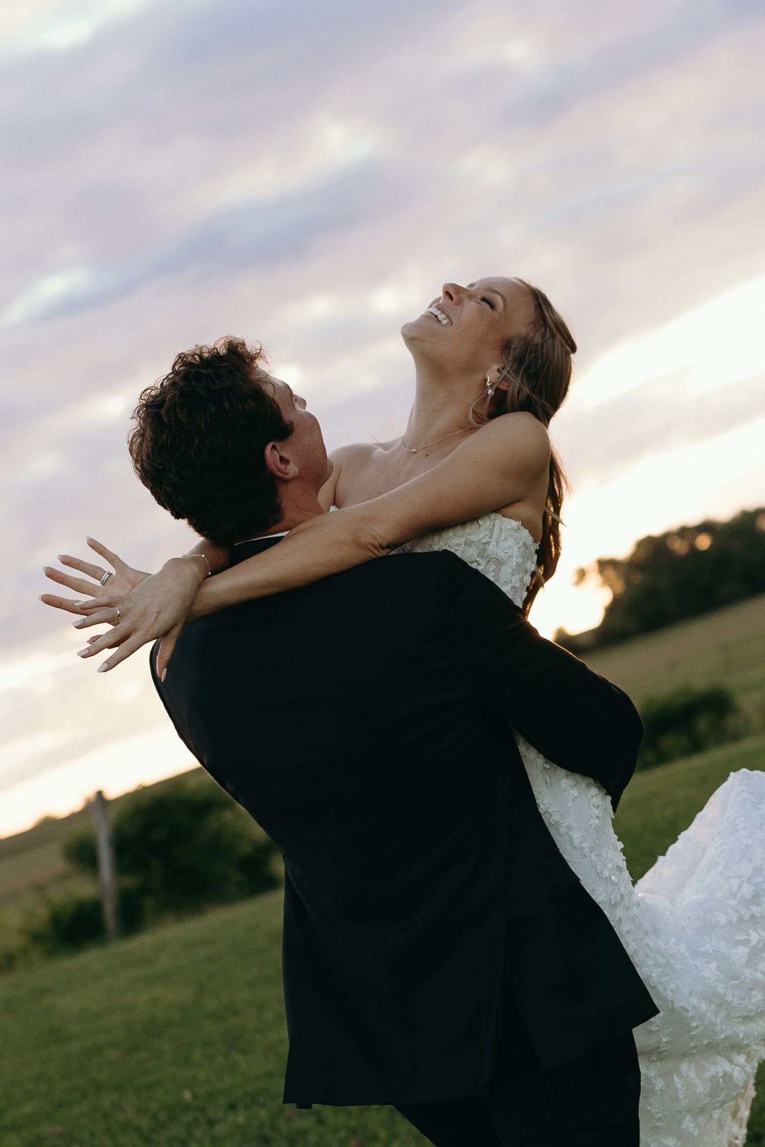 A couple in wedding attire embrace joyfully in a grassy field at sunset, creating a romantic silhouette against the sky.