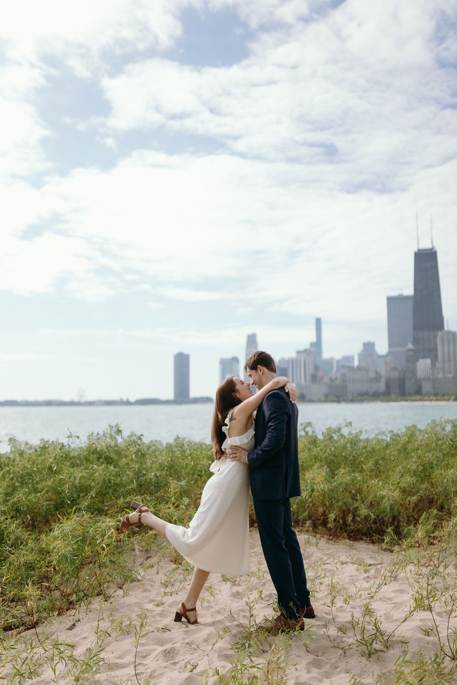 A romantic couple embraces on the sandy shore with Chicago's iconic skyline and Lake Michigan in the background.