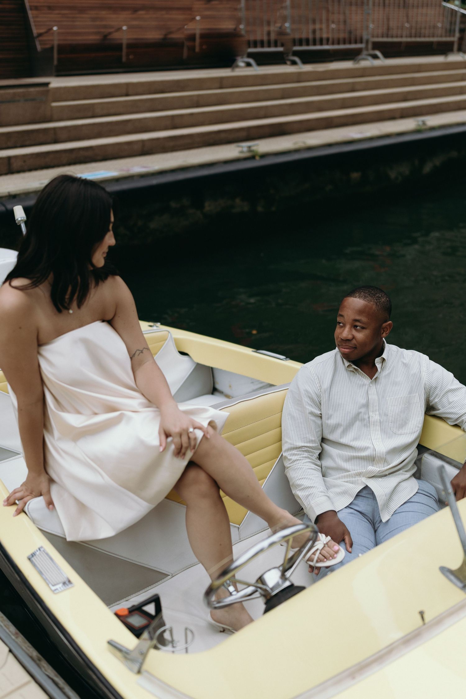 A couple enjoys a romantic boat ride, with a woman in a pink dress and a man in casual attire on a yellow boat.