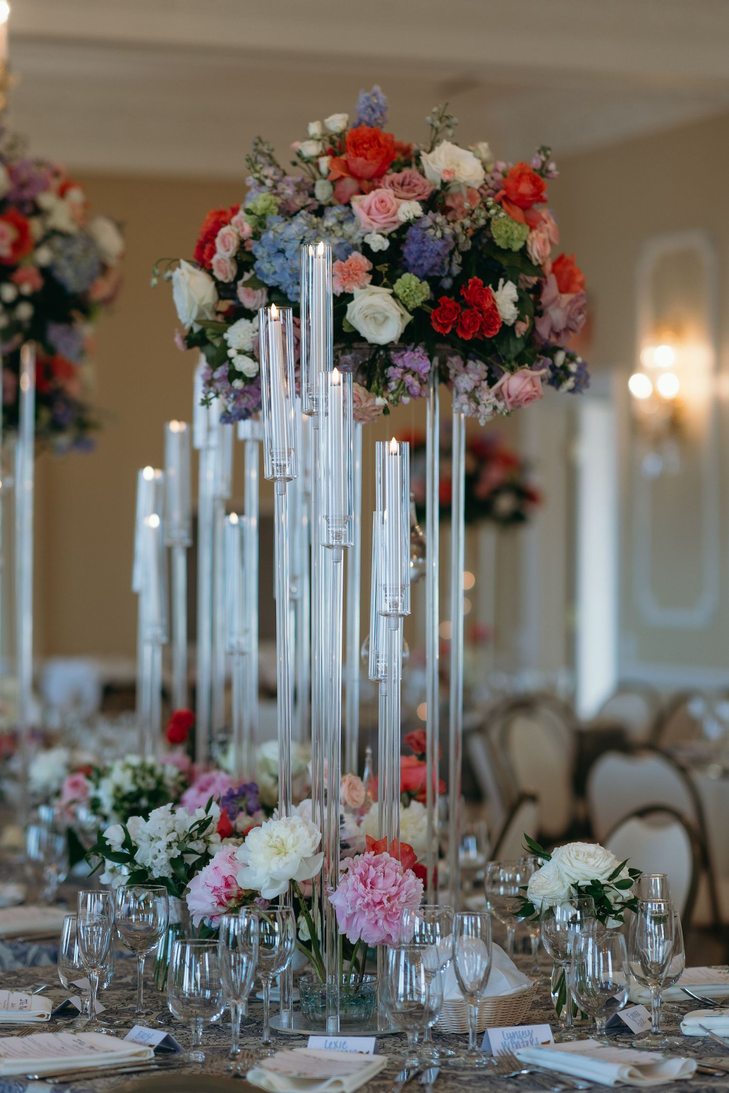 Elegant wedding centerpiece features tall crystal candlesticks topped with lush floral arrangements in pink, red and blue blooms.