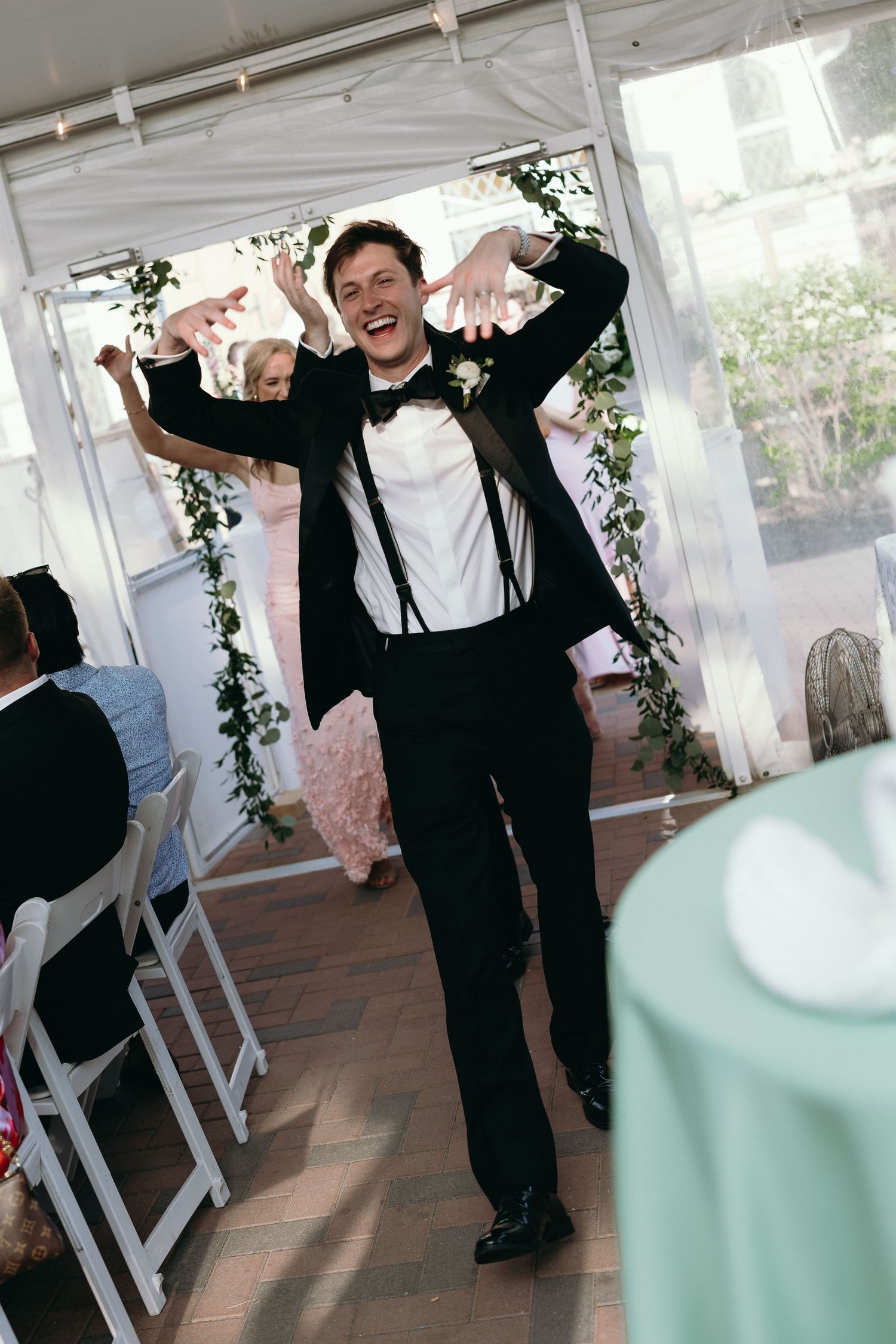 Groom in black tuxedo dancing joyfully at wedding reception, making fun gestures with hands raised.