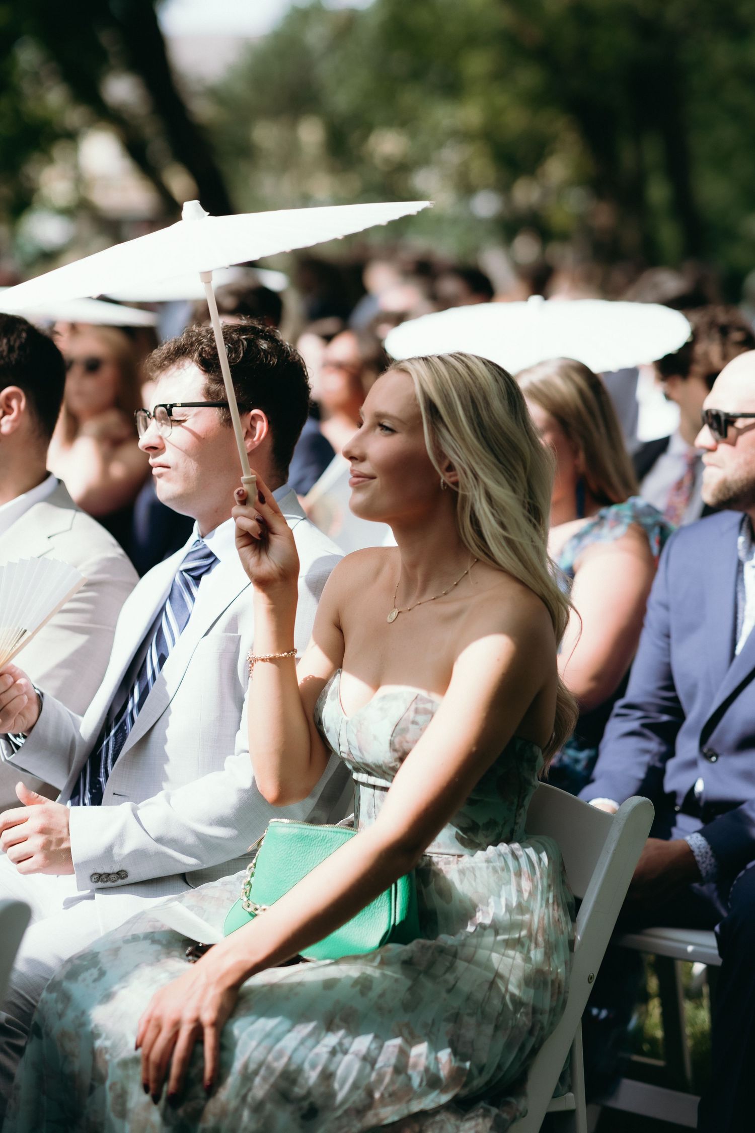 Elegant blonde woman in floral dress holding white parasol at outdoor wedding ceremony.