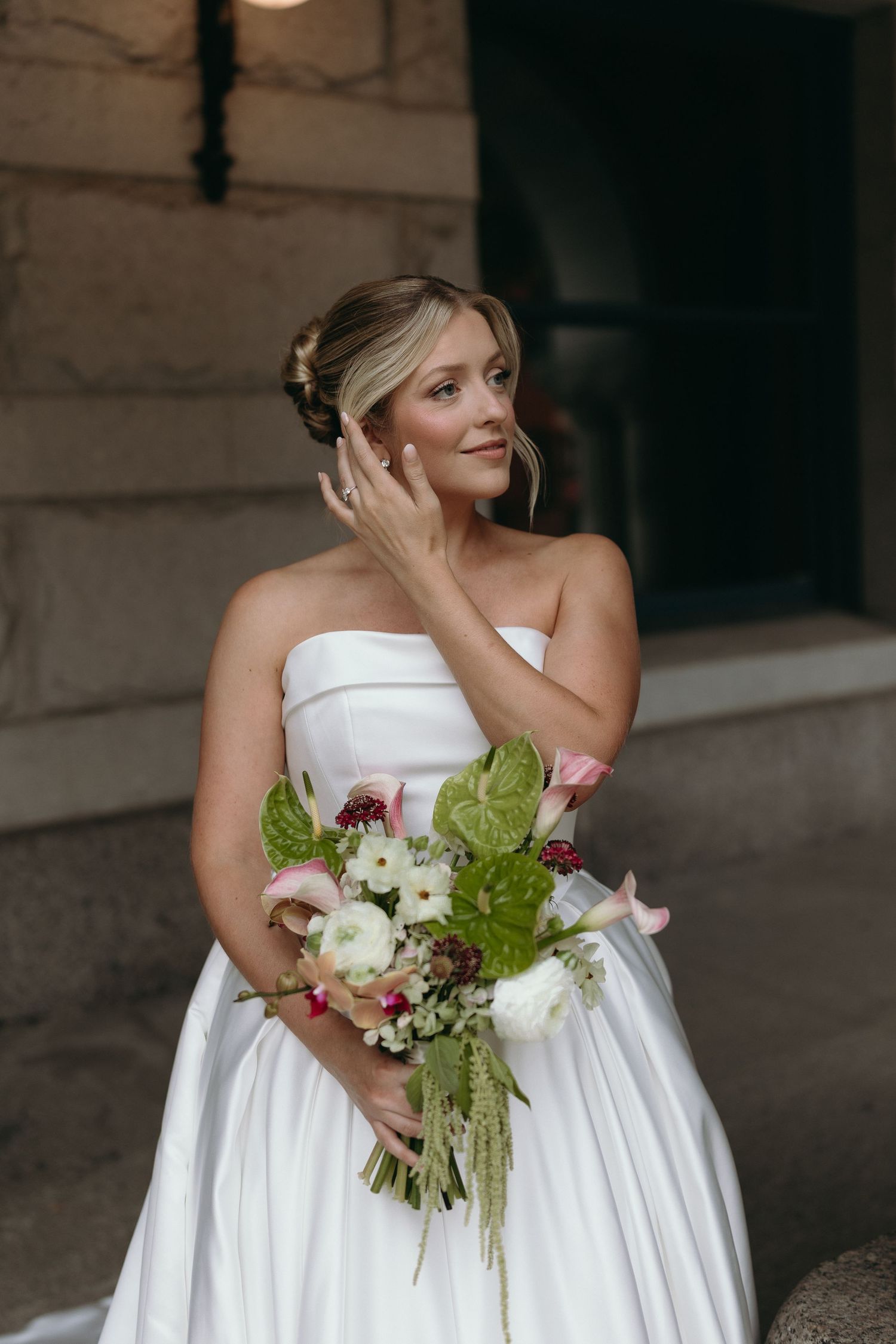 A bride in a strapless white wedding gown holds a bouquet of white flowers and greenery while posing outdoors.