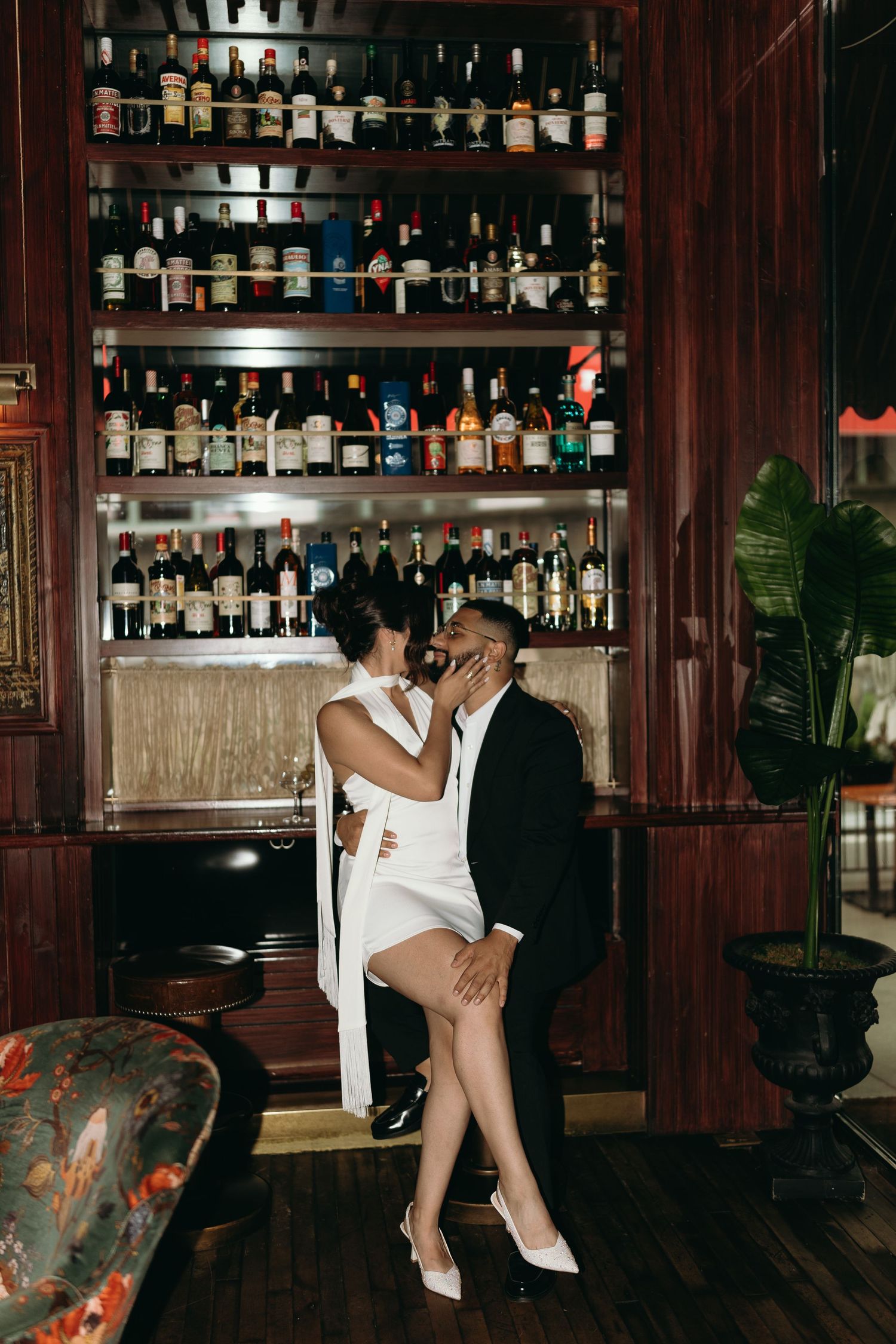 Couple in formal attire sharing intimate moment by vintage bar with extensive liquor collection.