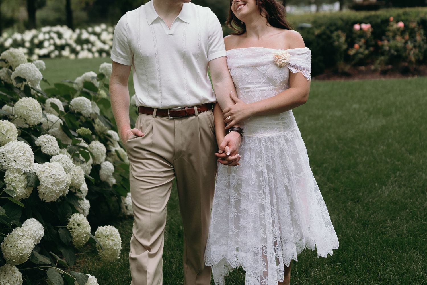 A couple embraces on a manicured lawn with white hydrangea bushes in a romantic outdoor garden setting.