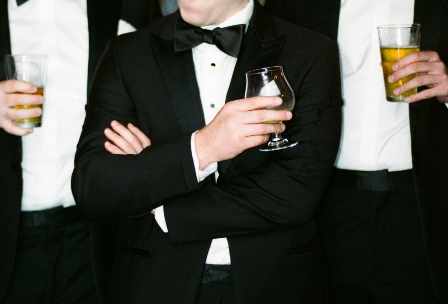 Men in black tuxedos with bow ties holding cocktail drinks at a formal wedding reception event.
