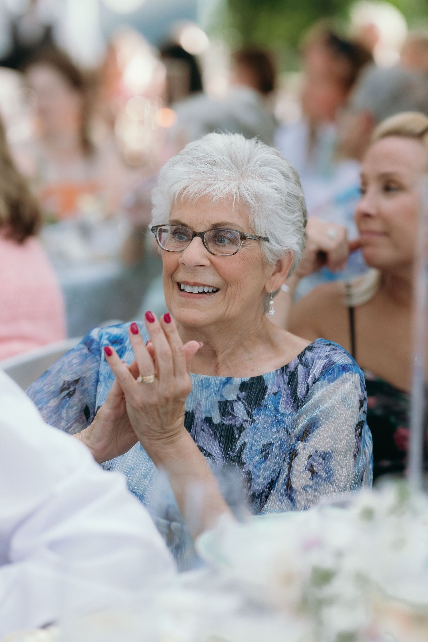 Happy elderly woman with white hair and red nails clapping and smiling at outdoor wedding reception.