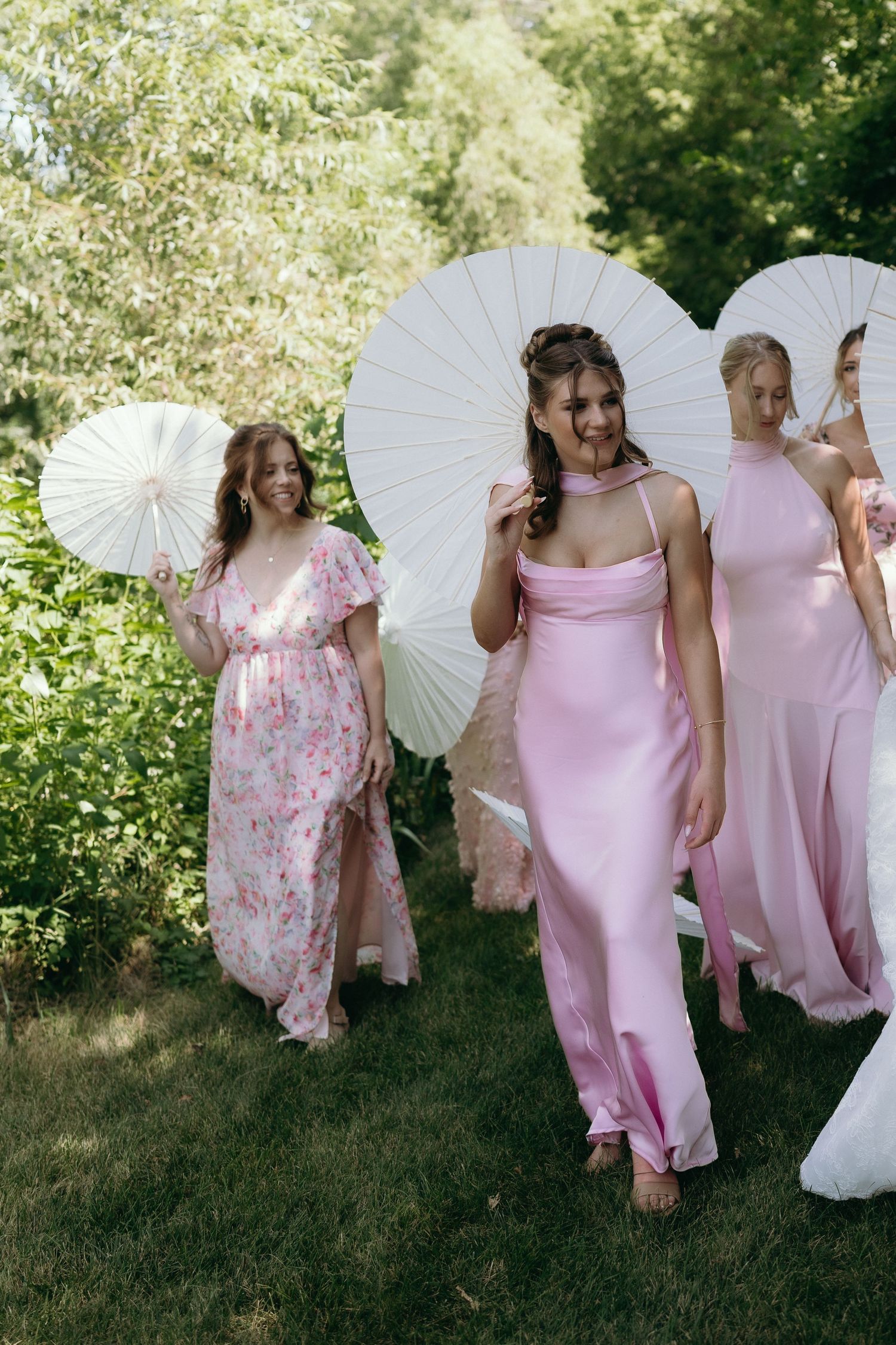 Bridesmaids in pink dresses carrying white parasols walk outdoors at a garden wedding ceremony.