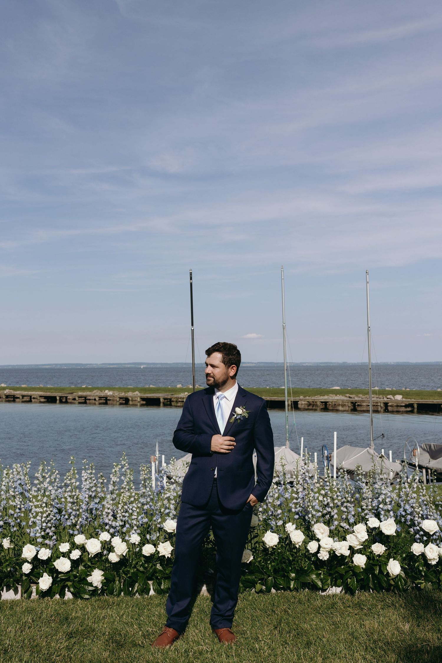 Formal portrait of person in navy suit standing in garden with white flowers and waterfront view behind on overcast day.