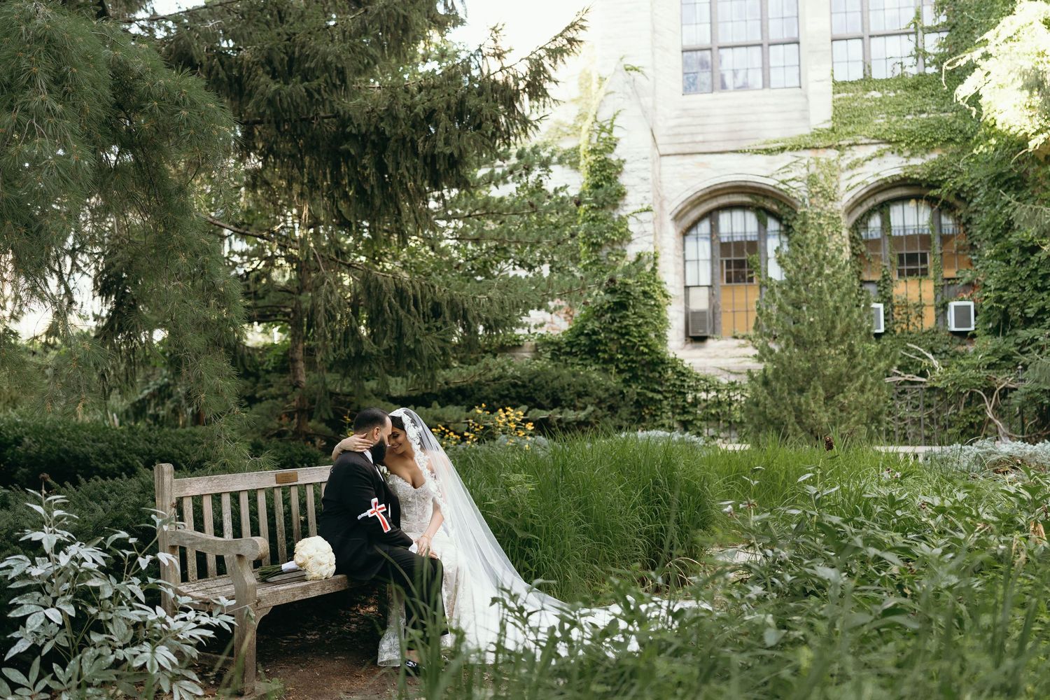 A wedding couple sits on a garden bench surrounded by lush greenery and architecture.