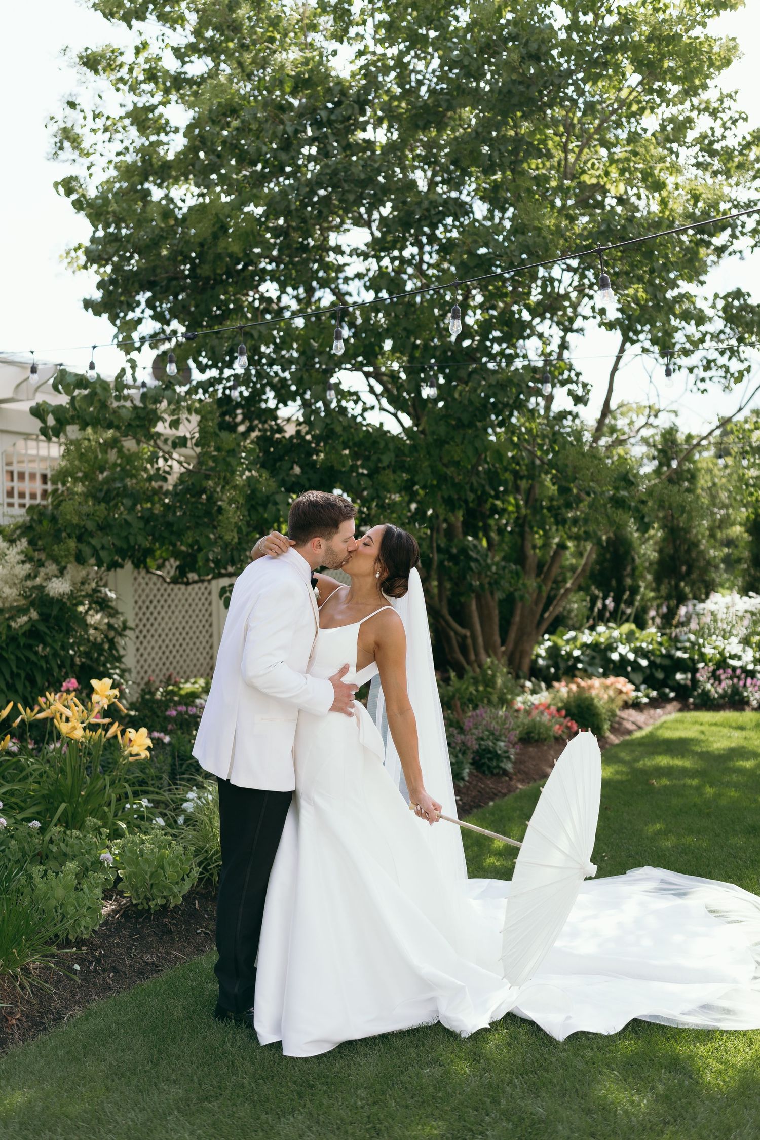 Wedding couple embraces on a green lawn with trees in the background.