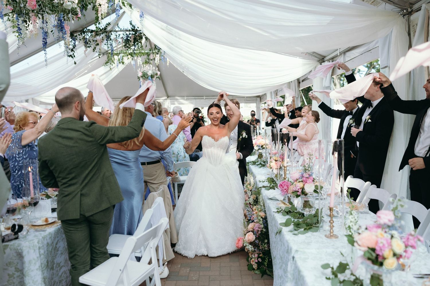 Bride in white gown makes grand entrance at outdoor wedding reception as guests cheer under a white tent.