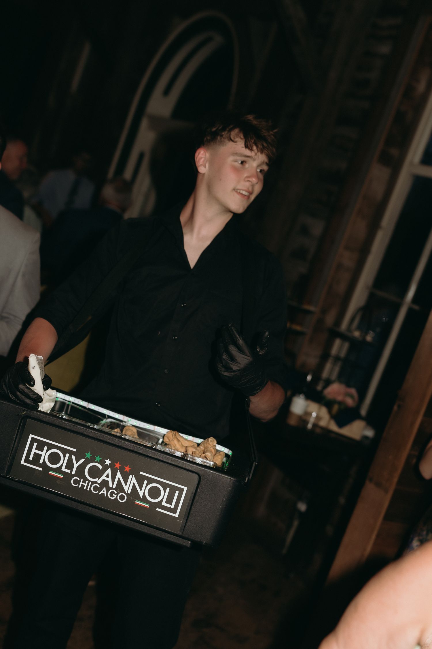 Server in black uniform carrying Holy Cannoli Chicago dessert tray at dark evening event.