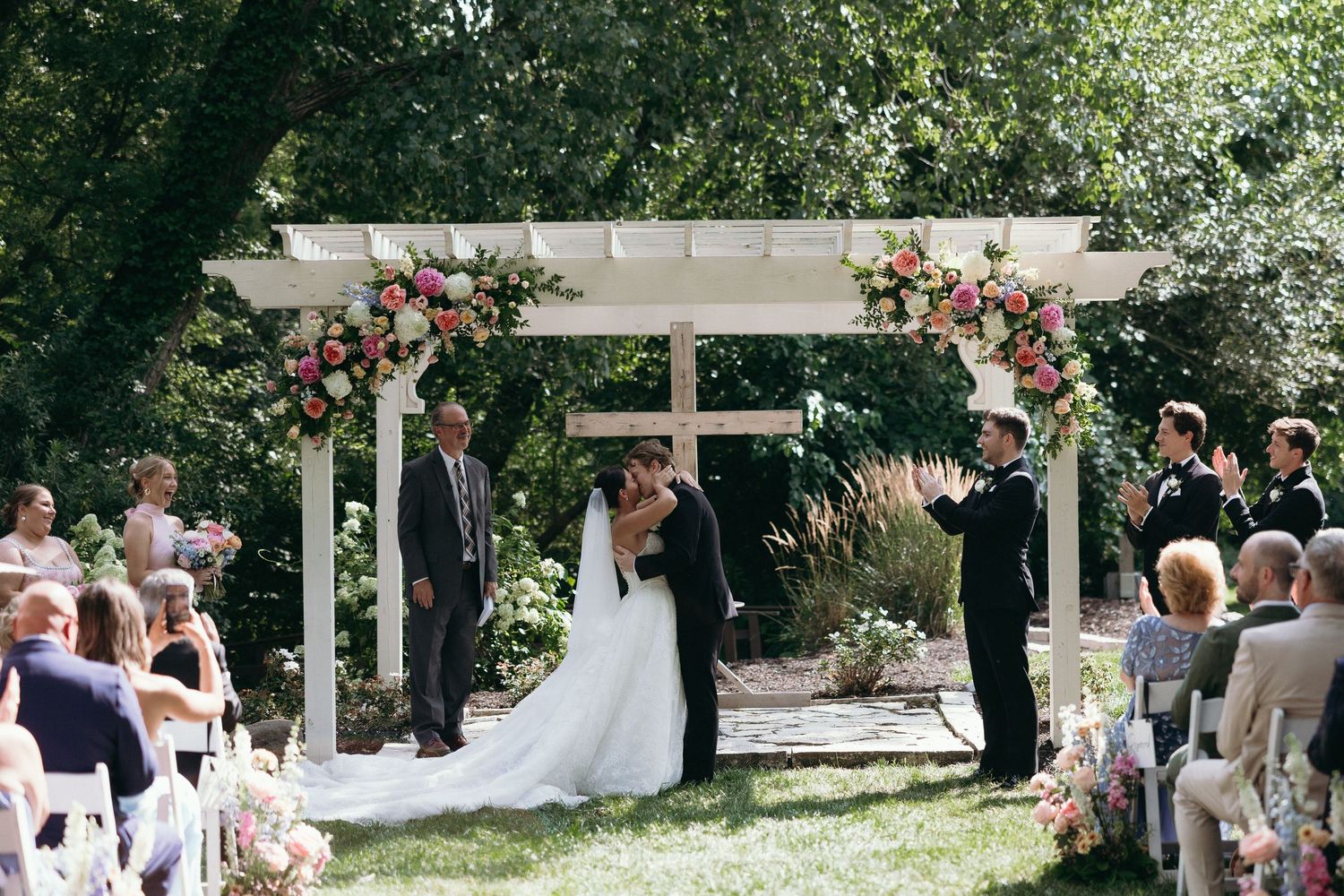 Bride and groom share first kiss at outdoor wedding ceremony under floral arch with guests seated on both sides.