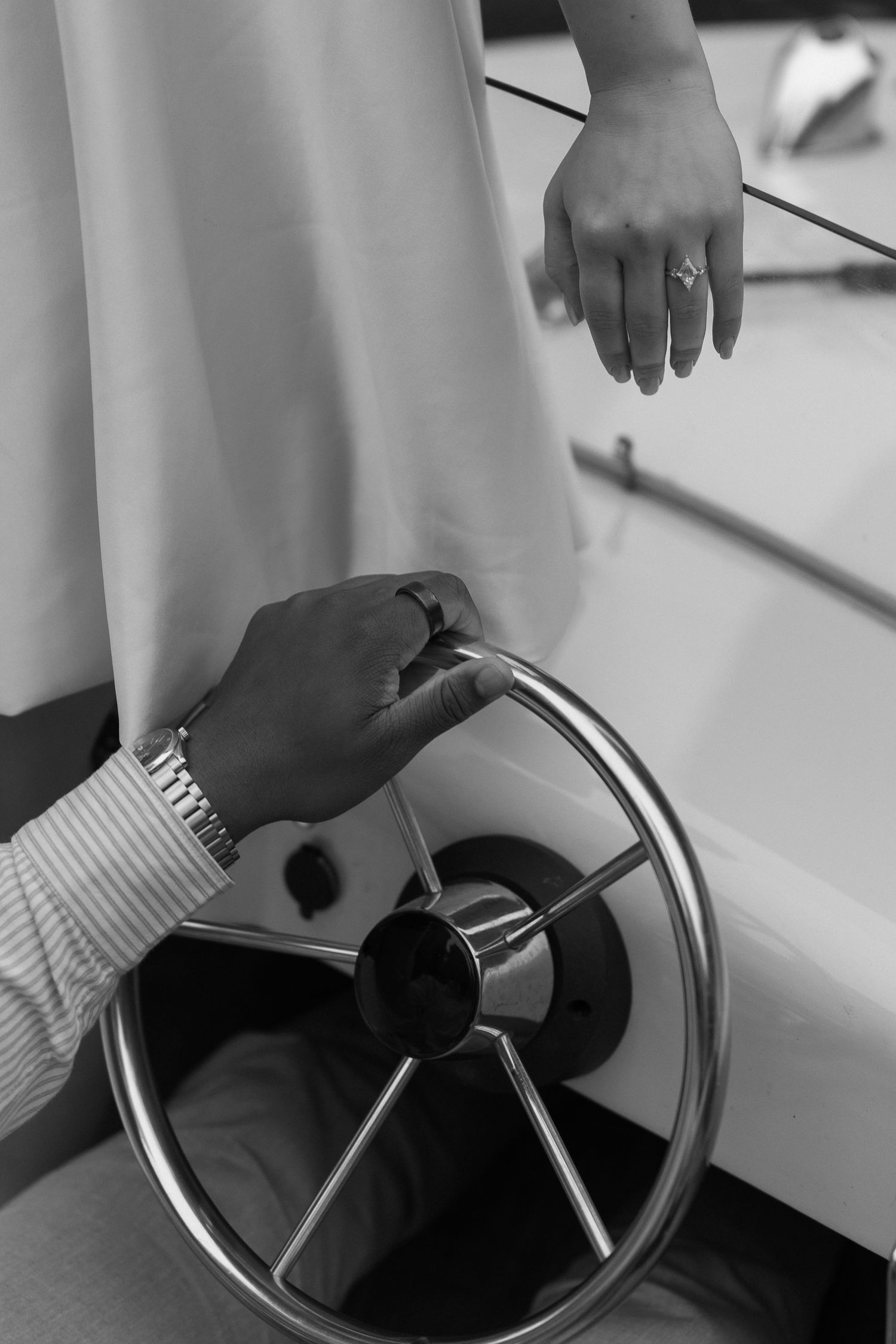 Black and white photo of a couple on a boat, groom steering wheel, bride wearing engagement ring nearby.