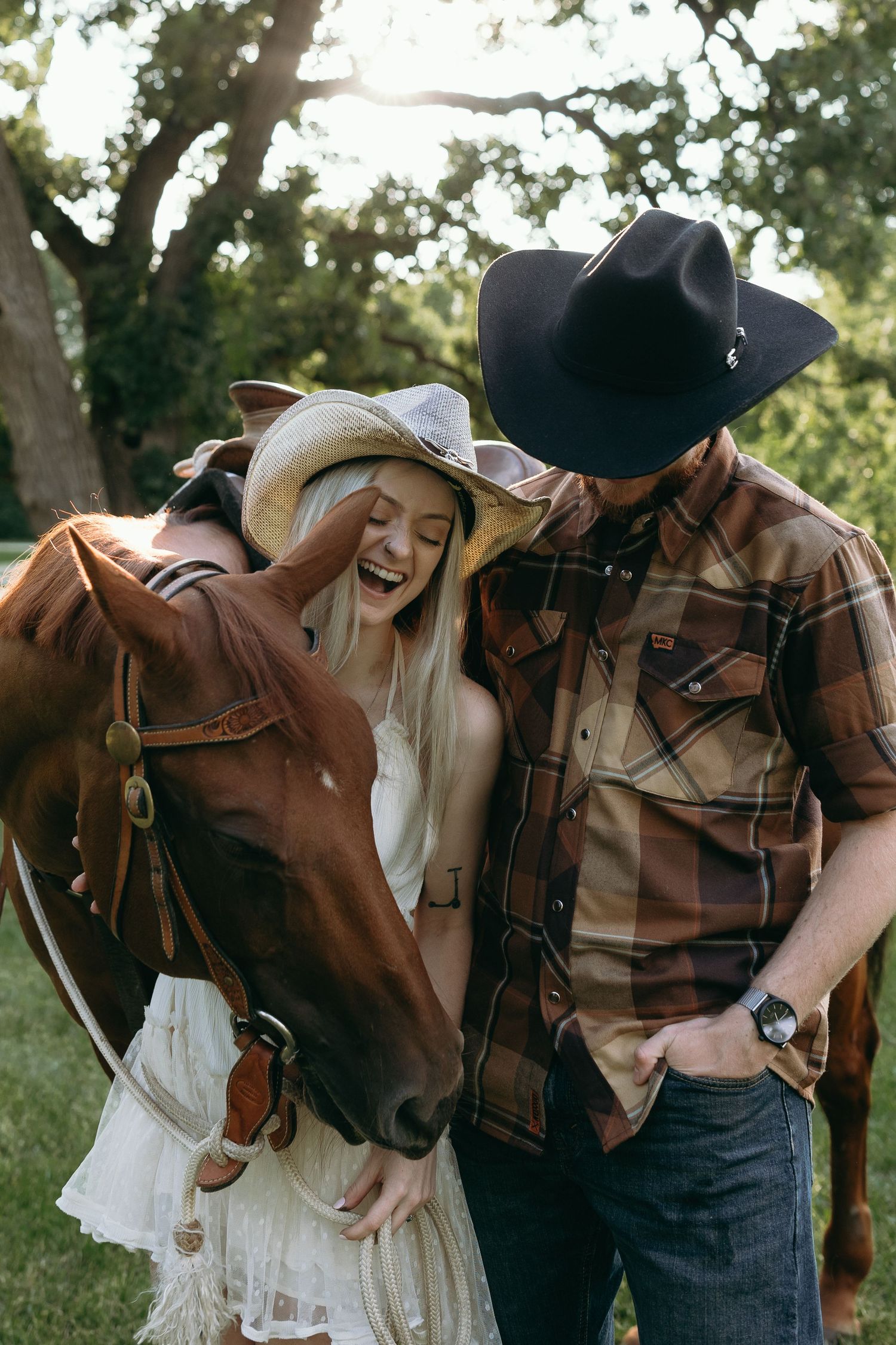 A couple in western wear share a joyful moment with their brown horse in a sunlit outdoor setting.