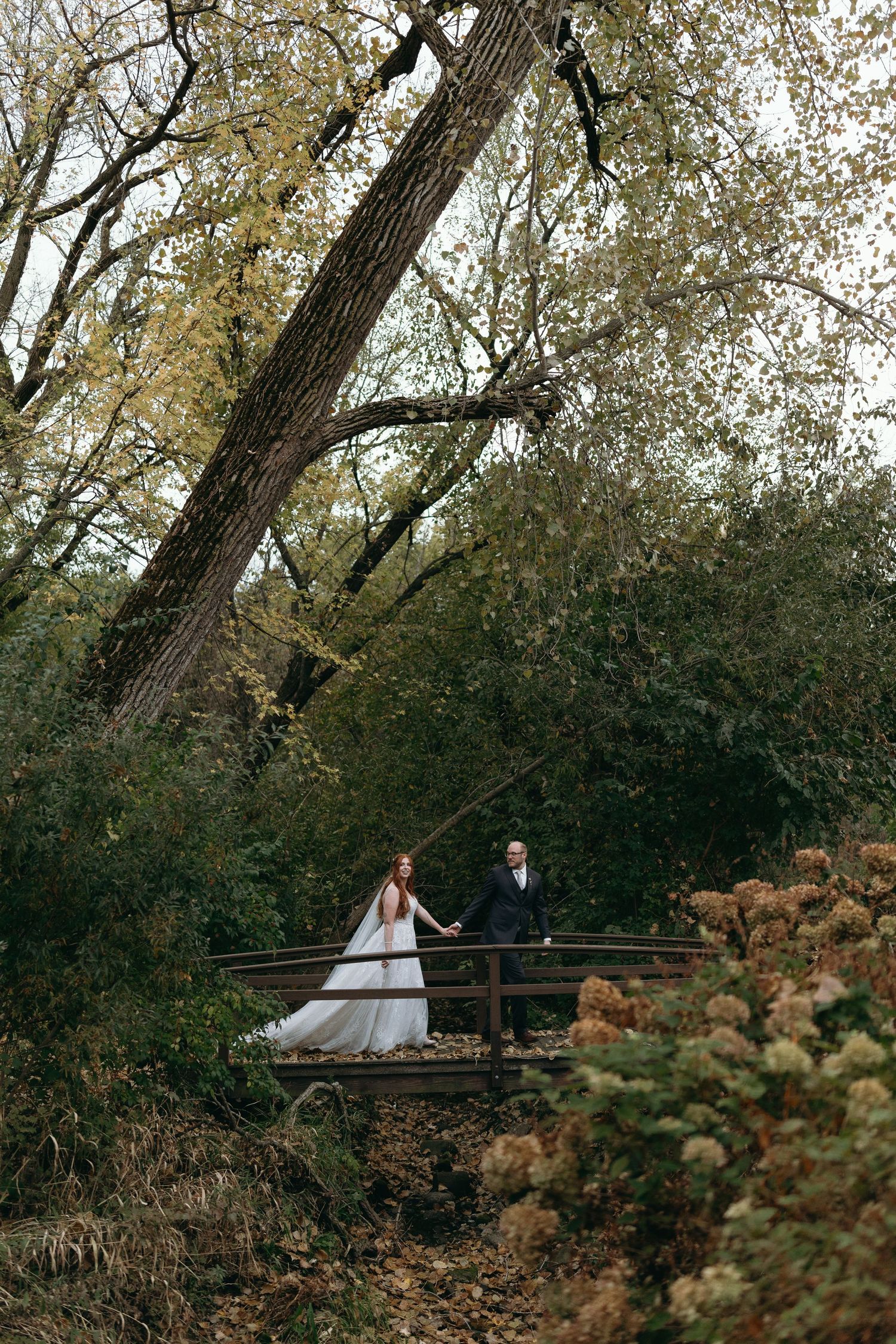 A couple walks together across a wooden bridge surrounded by autumn trees and golden foliage in a romantic forest setting.