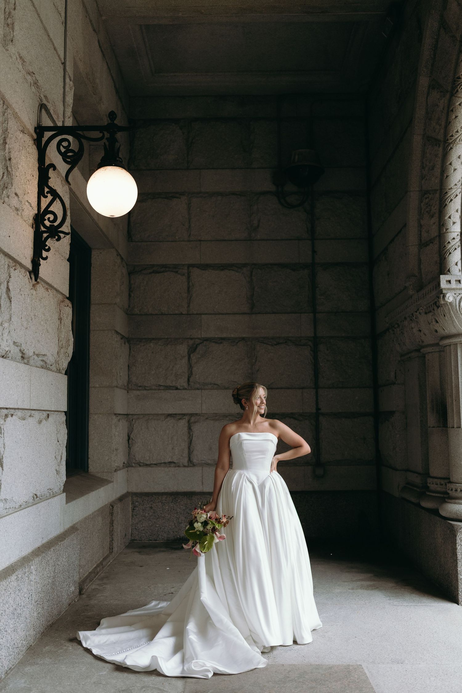A bride in a flowing white wedding gown poses elegantly in a rustic stone hallway.