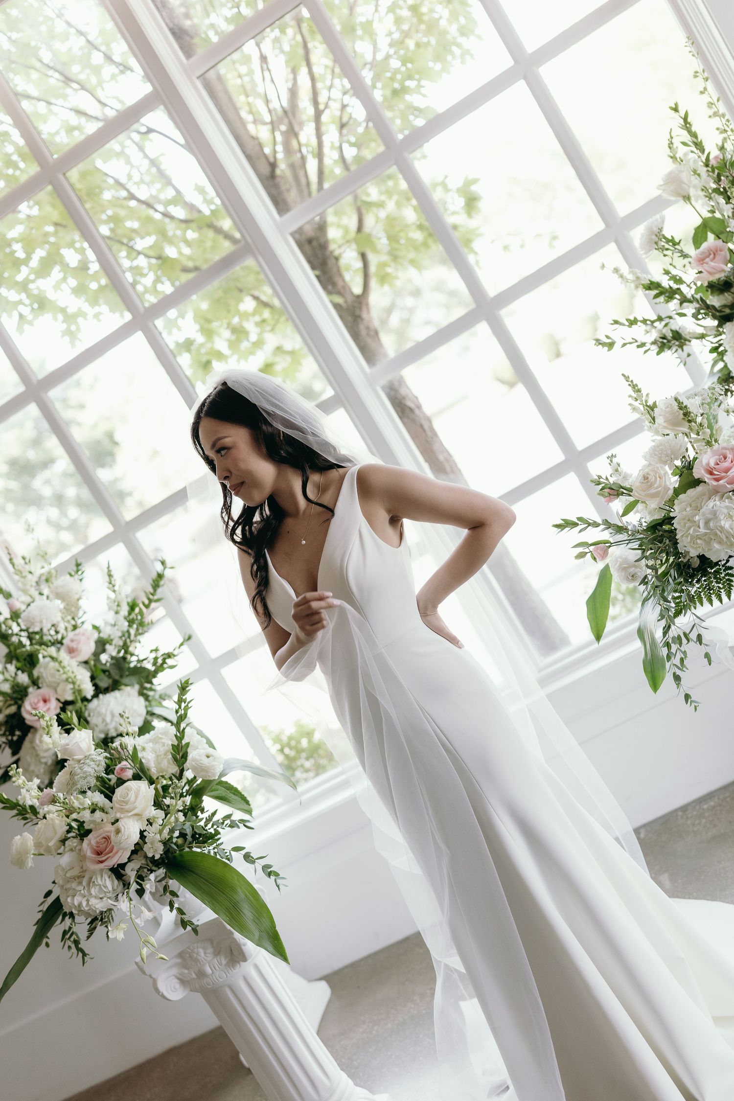 Elegant bride in flowing white dress poses gracefully in sunlit conservatory with floral arrangements.