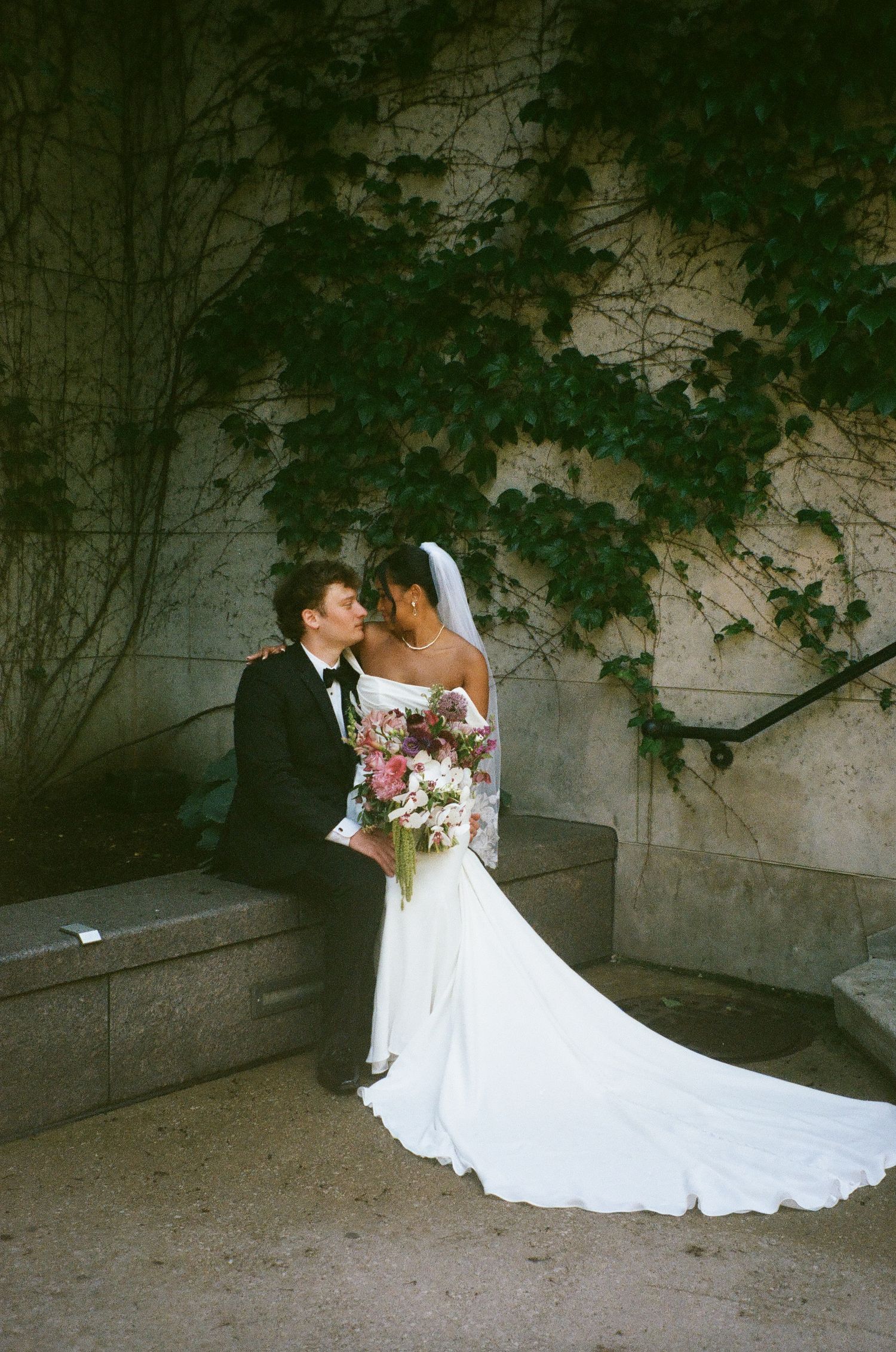 Bride and groom share a romantic moment seated on stone steps, surrounded by ivy, bride holding a lush floral bouquet.