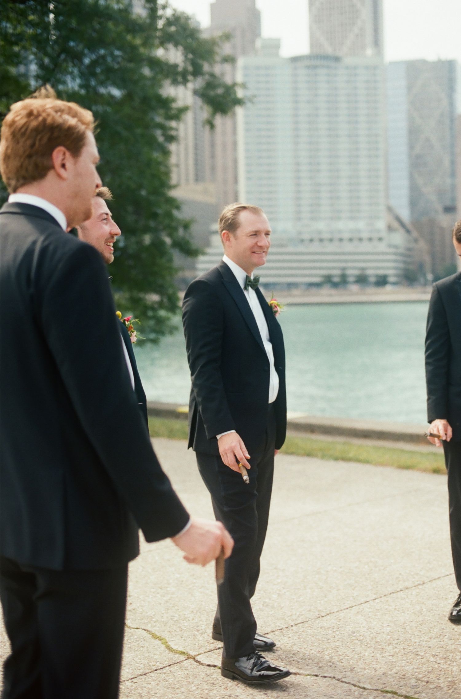 Groomsmen in black tuxedos laughing outdoors near a river with city skyline backdrop on a wedding day.