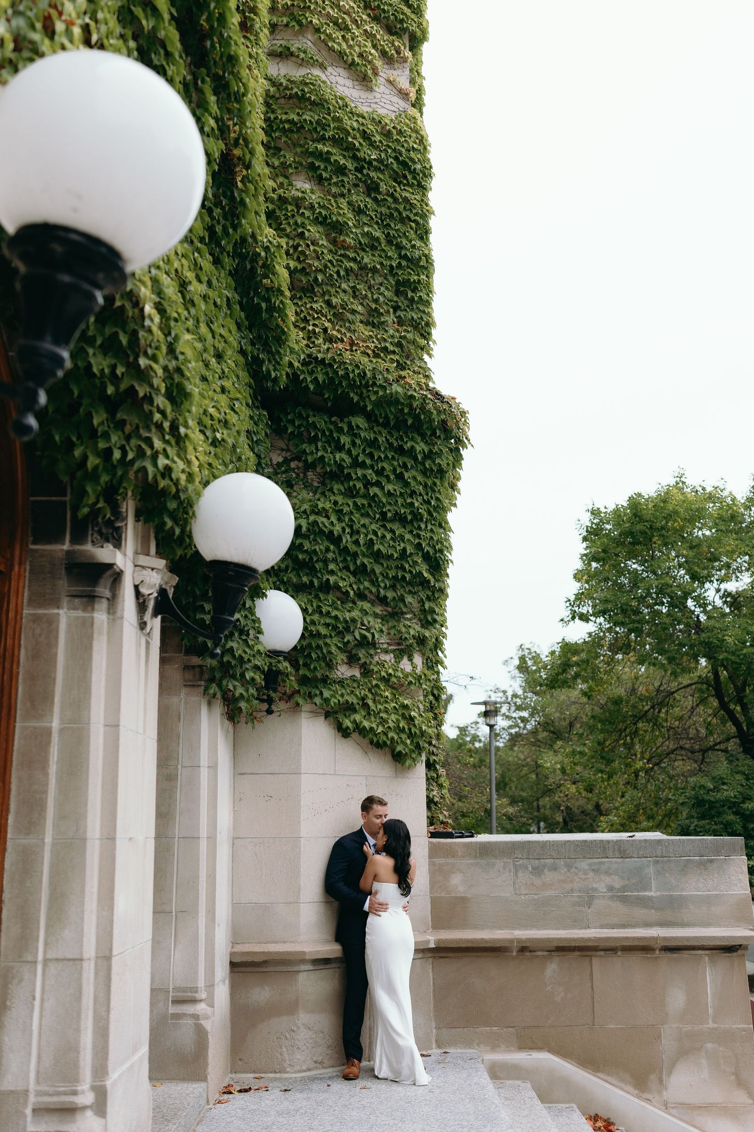 A romantic embrace on stone steps beside an ivy-covered wall with white globe lights.