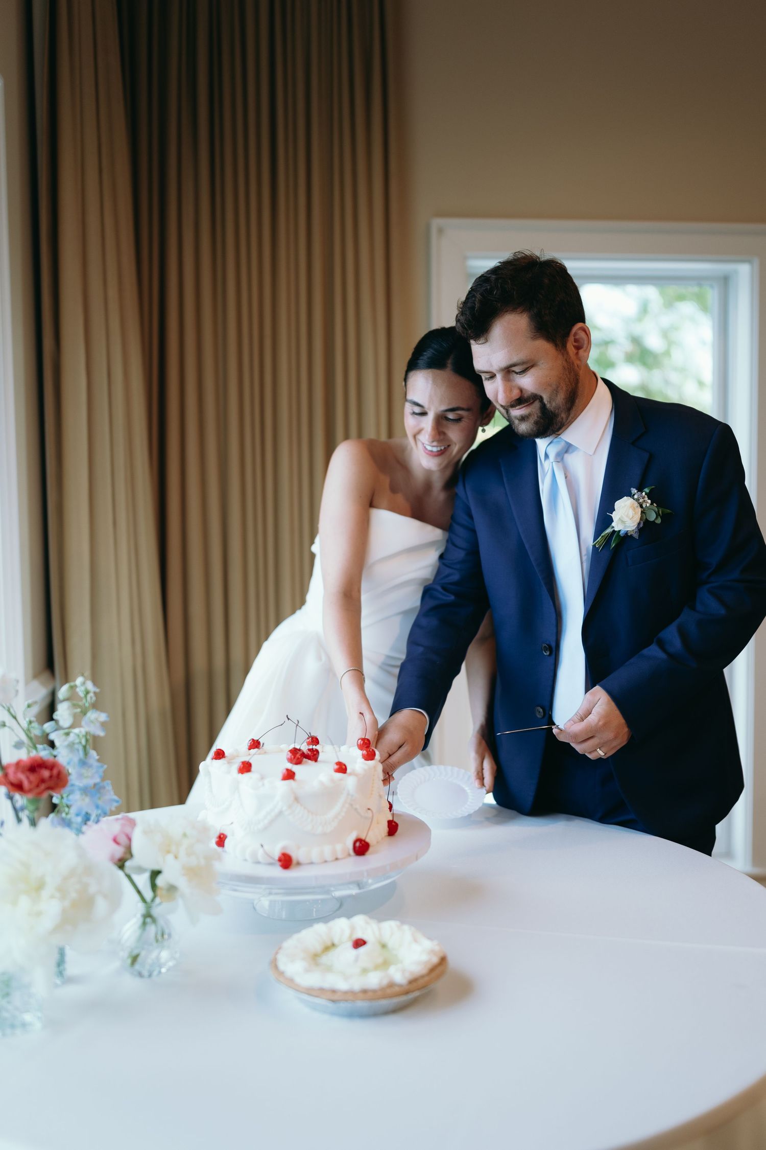 Newlywed couple shares sweet moments while cutting their white wedding cake adorned with red flower petals.