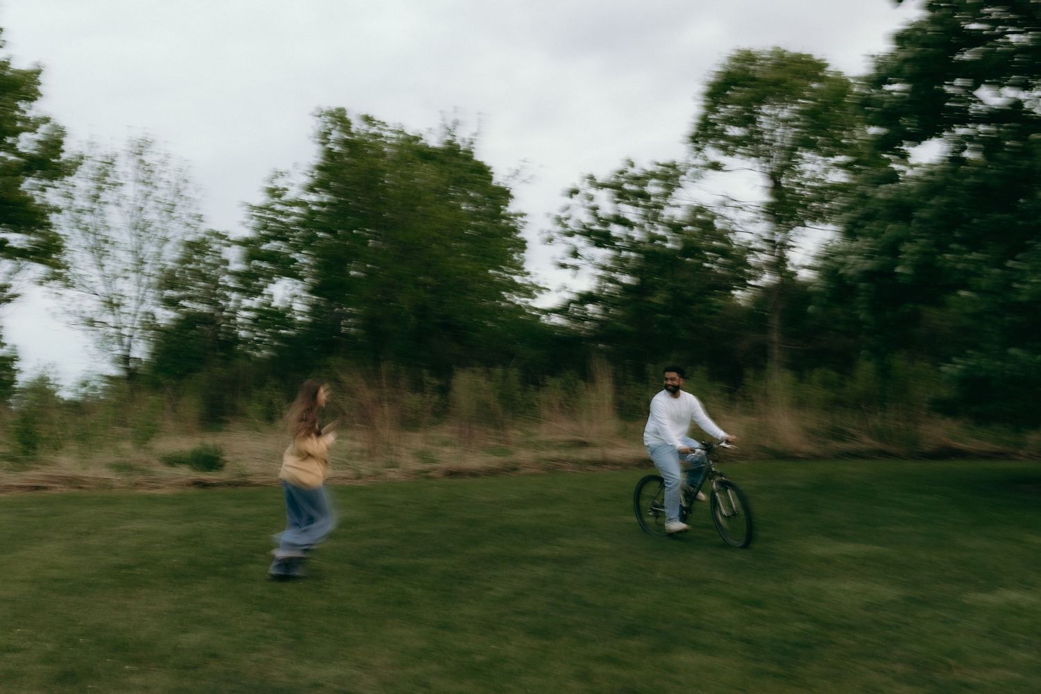 Chicago engagement photographer A person riding a bicycle and another running on a lush green field surrounded by trees on a cloudy day.