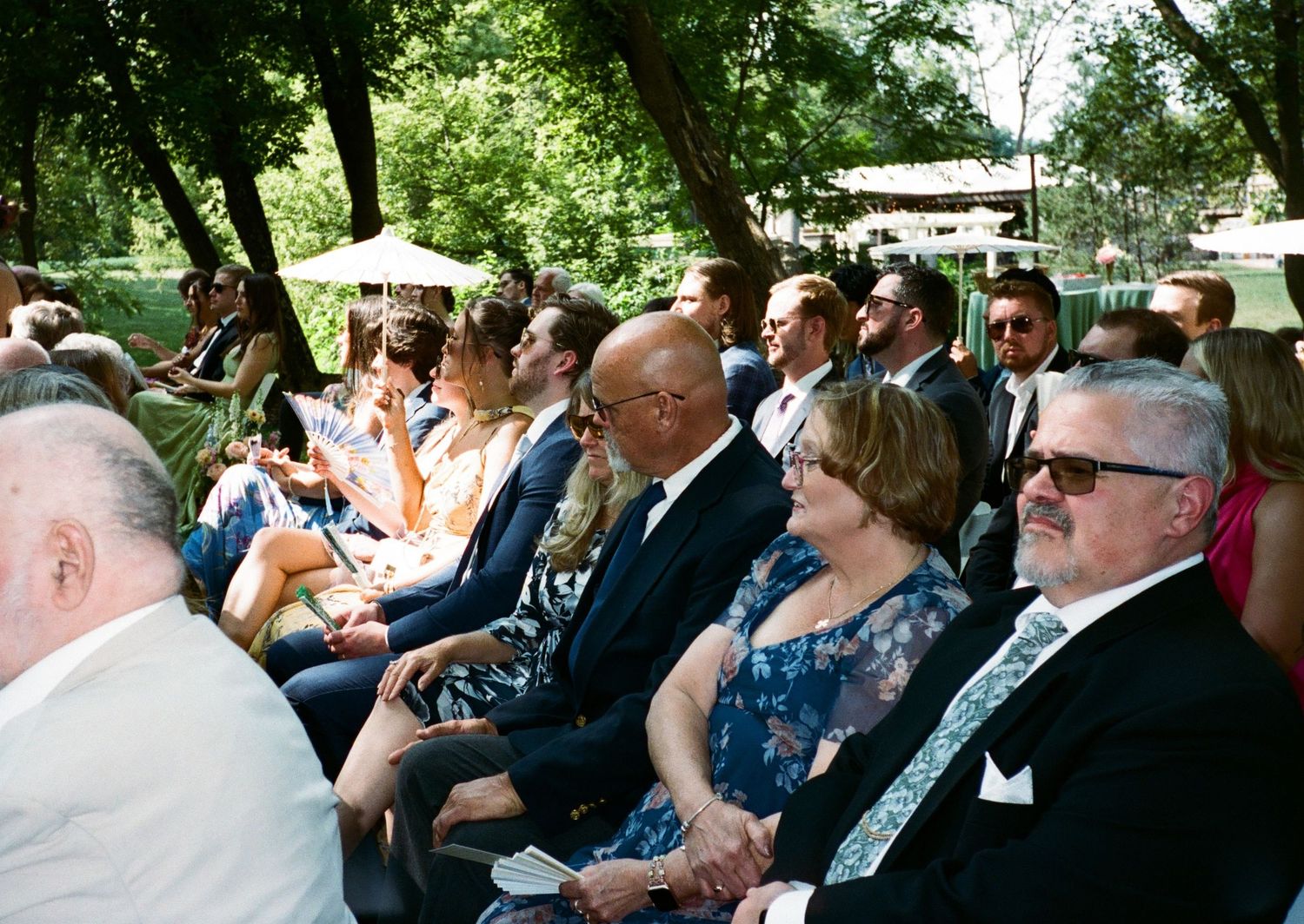 Guests seated outdoors at a wedding ceremony, dressed in formal attire, enjoying the celebration under shaded trees.