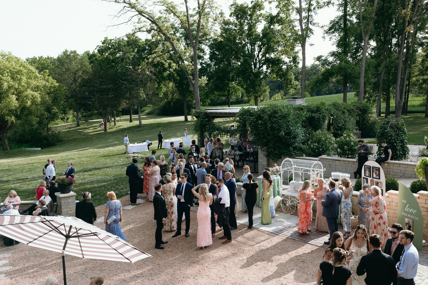Aerial view of wedding cocktail hour guests mingling outdoors on a sunny day surrounded by lush green trees.