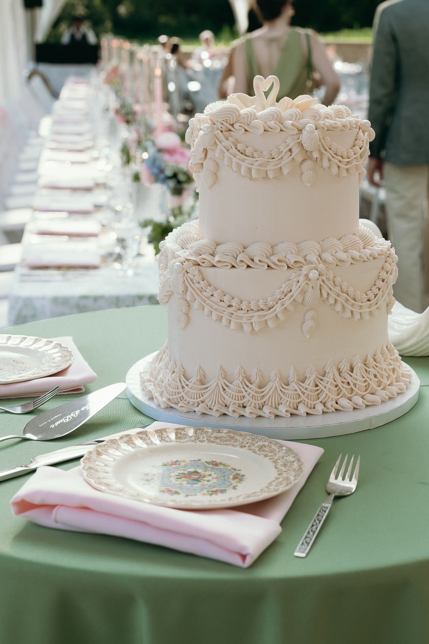 Elegant two-tier white wedding cake with intricate piped frosting details displayed on a green tablecloth at an outdoor reception.