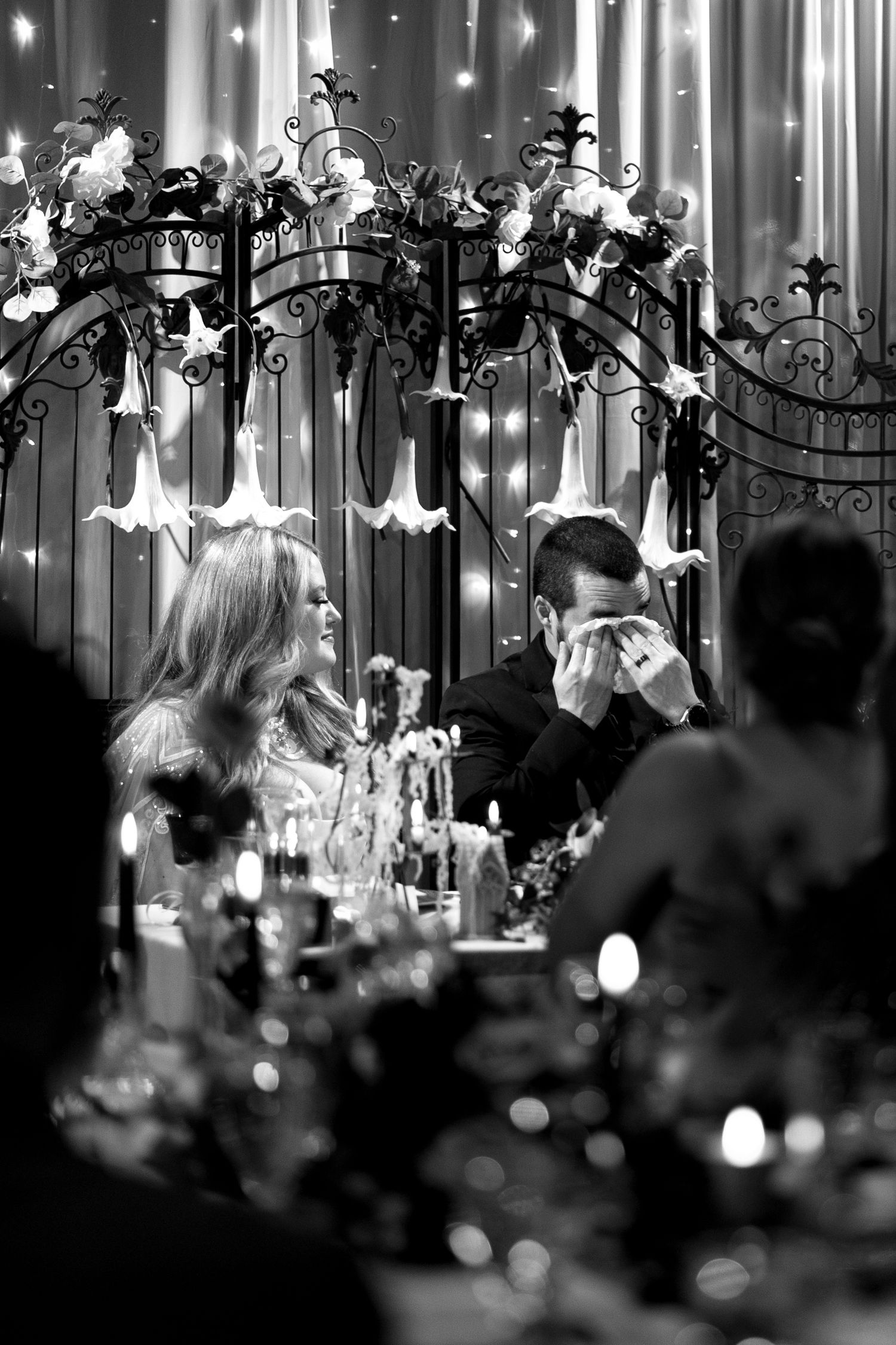 Black and white image of wedding reception under ornate floral arch with twinkling lights.