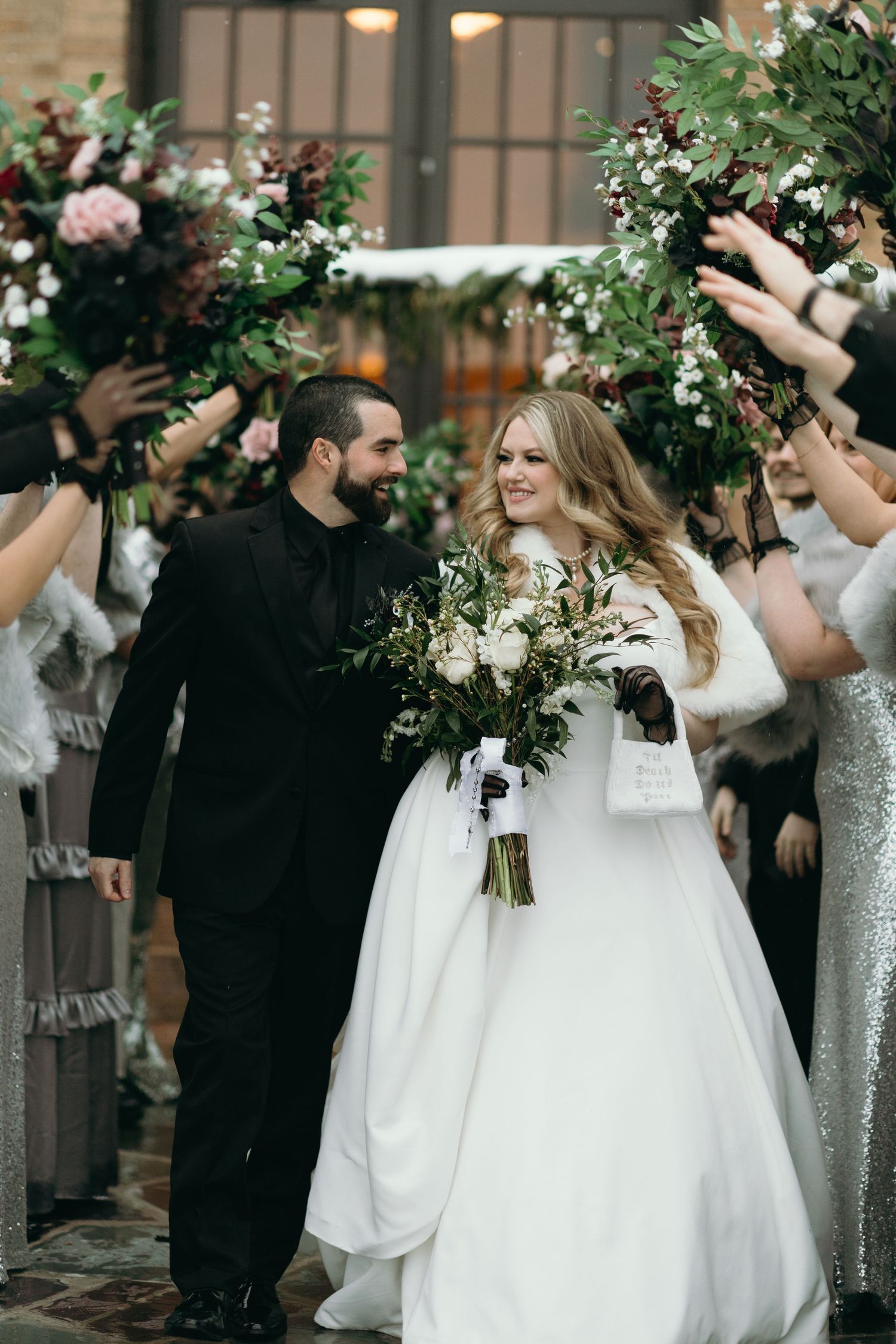 Two people walk beneath a floral wedding arch surrounded by guests with raised hands.