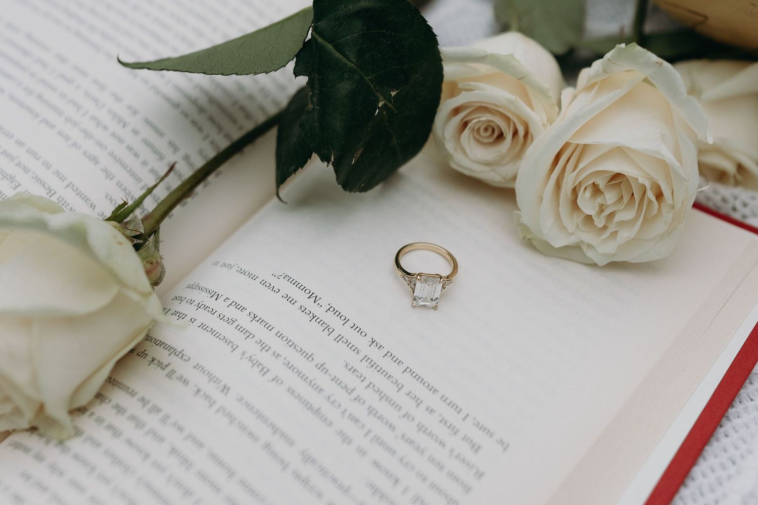 White roses and golden wedding rings rest atop open book pages in a romantic flat lay composition.