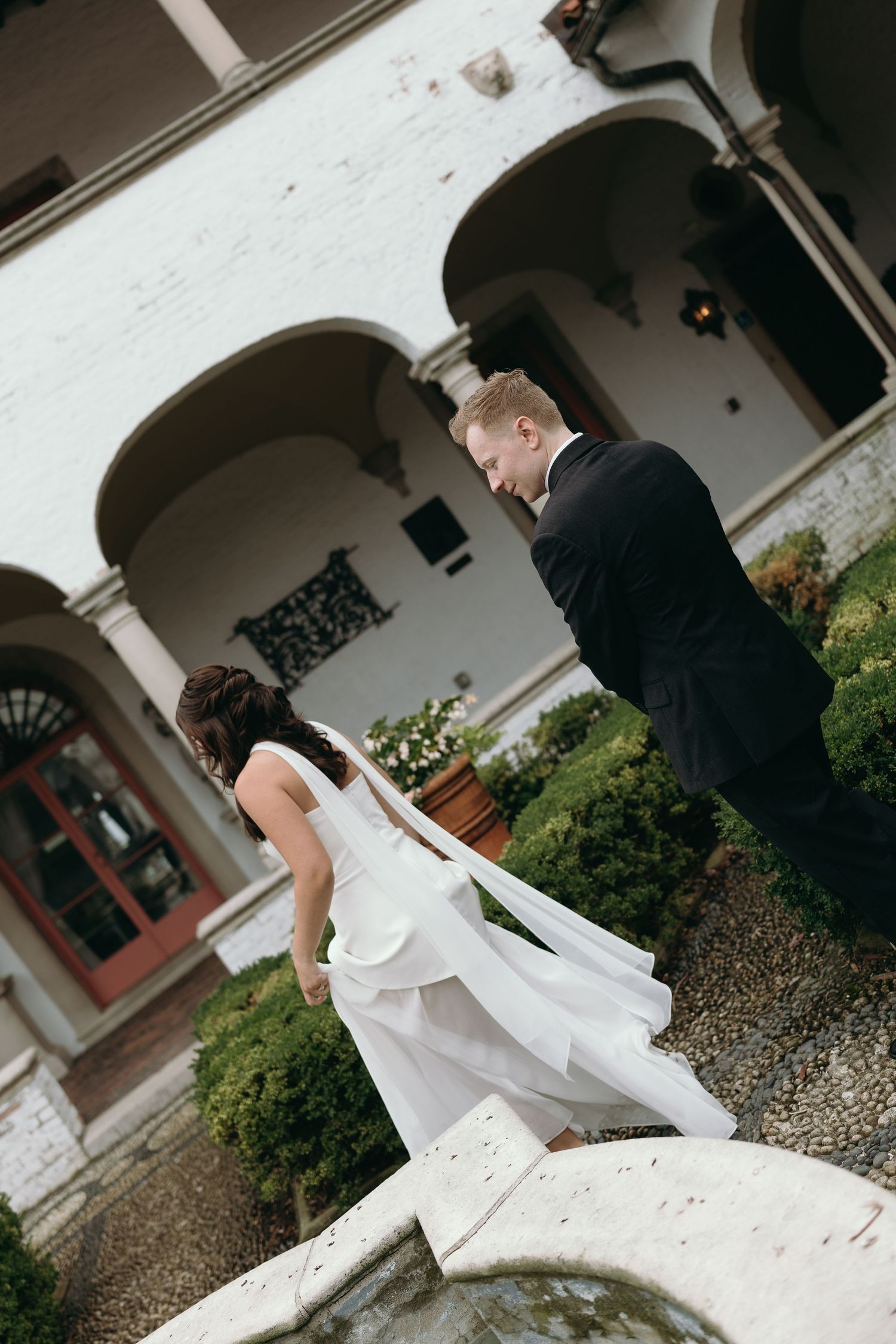 A couple dressed in formal attire dances together in an outdoor courtyard with white arches and greenery visible.