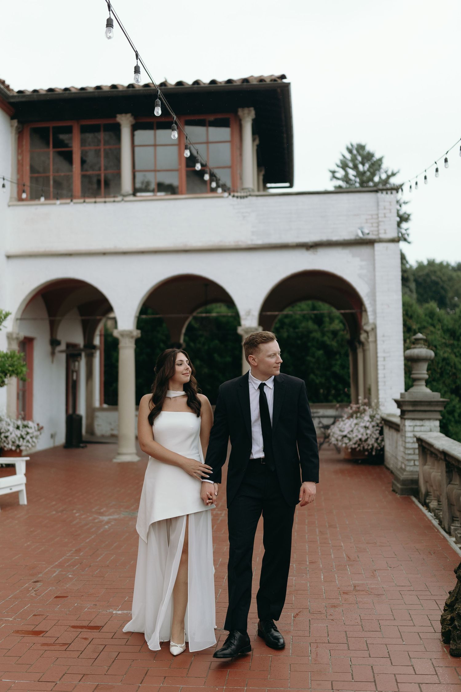 Elegant couple in formal attire stands on terracotta tile patio at Mediterranean-style venue with white arches.