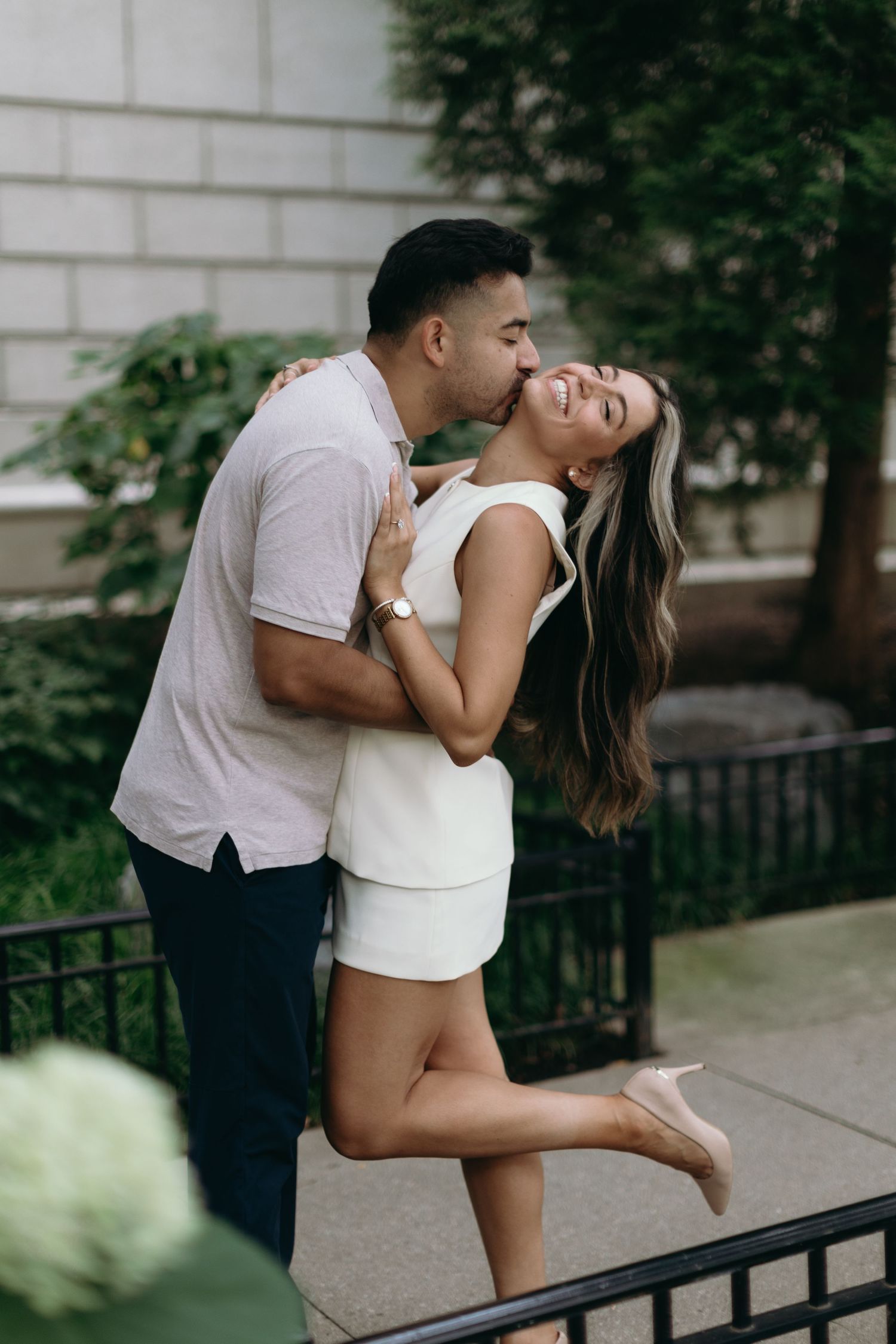 A romantic couple shares a passionate embrace and kiss while leaning against an outdoor railing in an urban setting.
