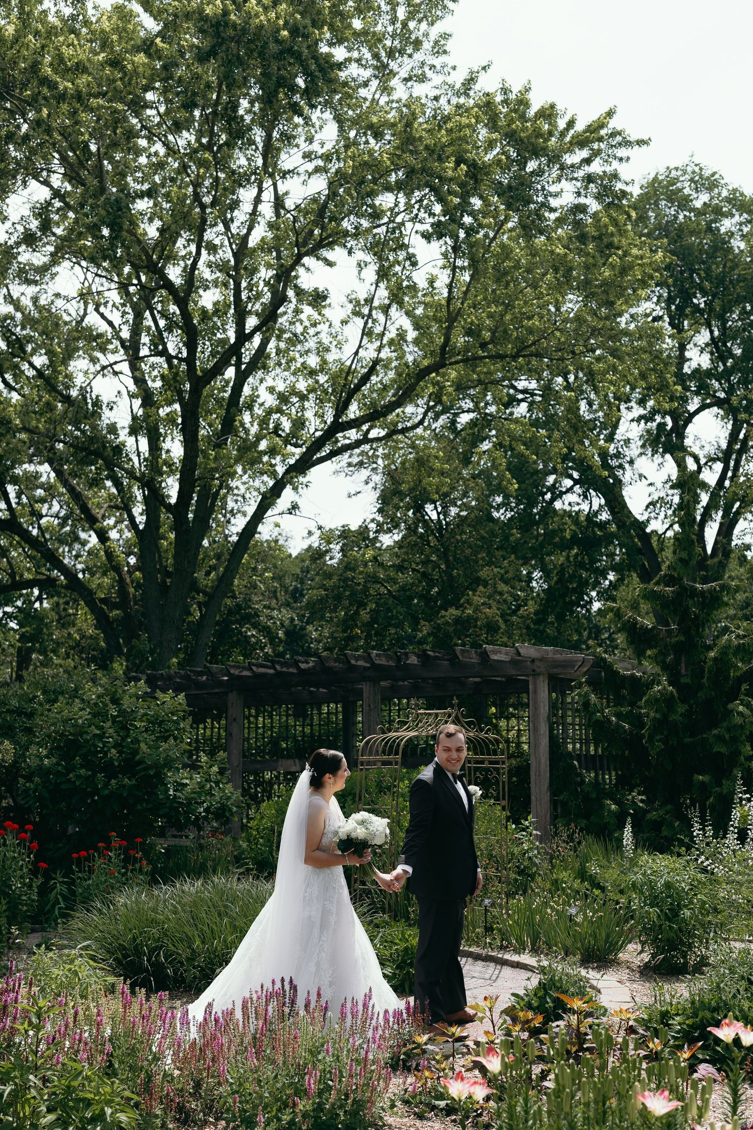 Wedding couple standing in garden with purple flowers and wooden pergola.