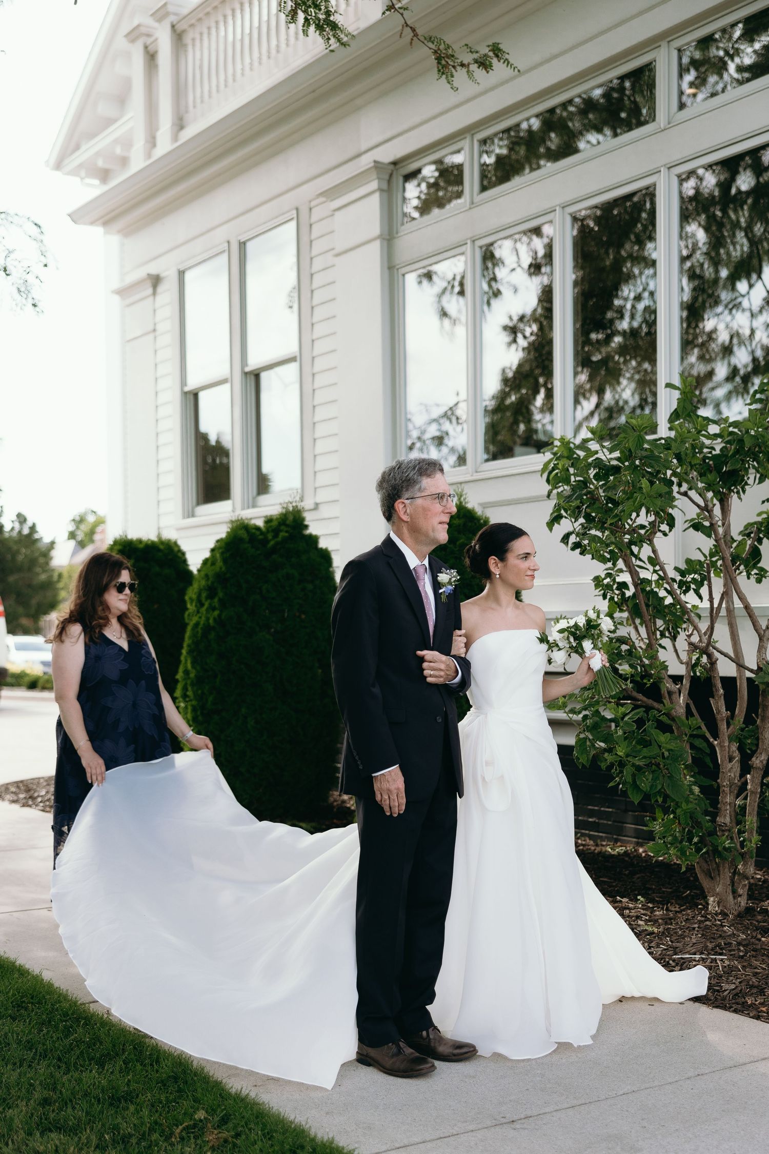 Wedding party member helps hold a long white bridal gown train outside a classic white house with elegant windows.