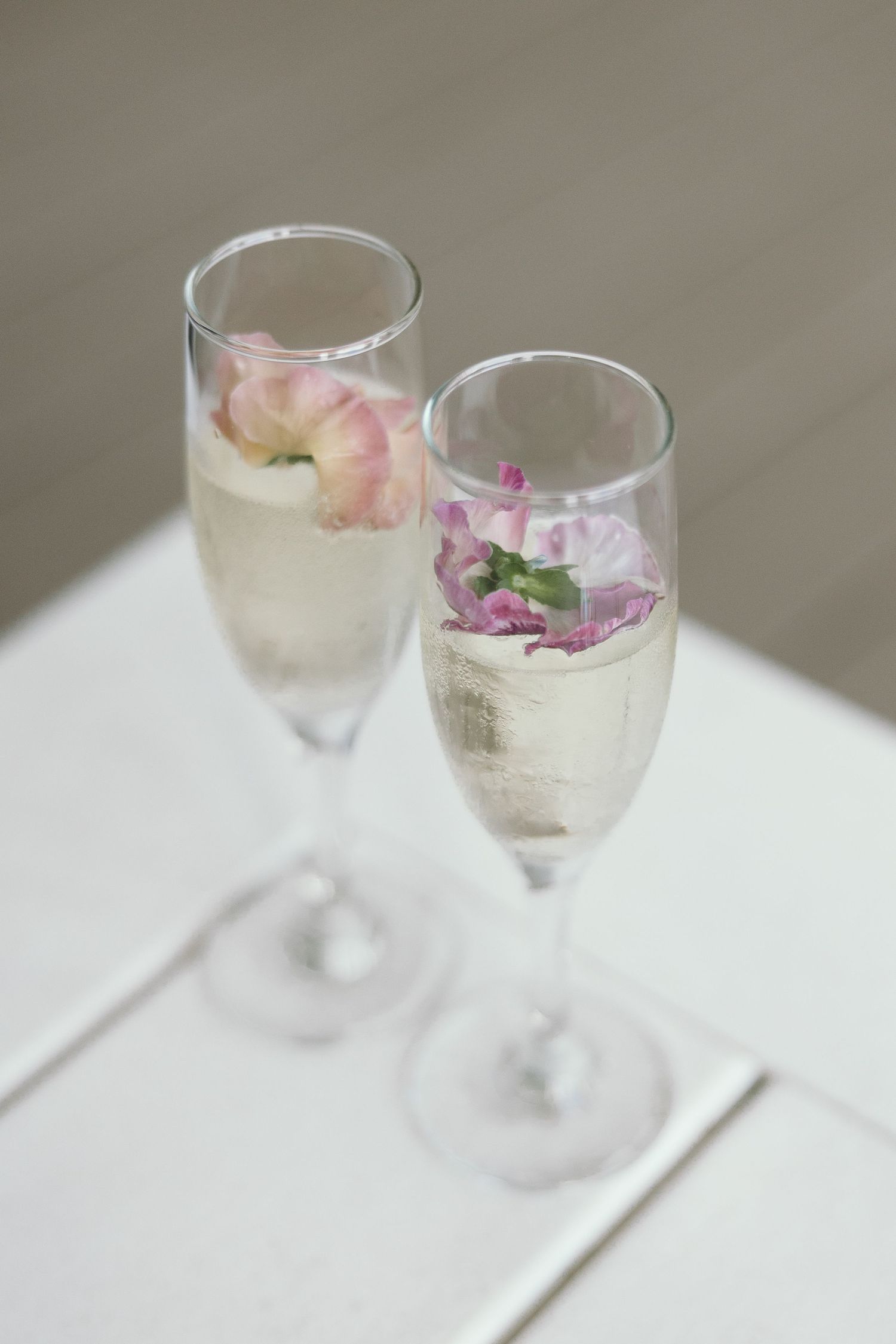 Two champagne flutes filled with sparkling wine and garnished with delicate pink and purple flower petals. Wedding Signature drink.