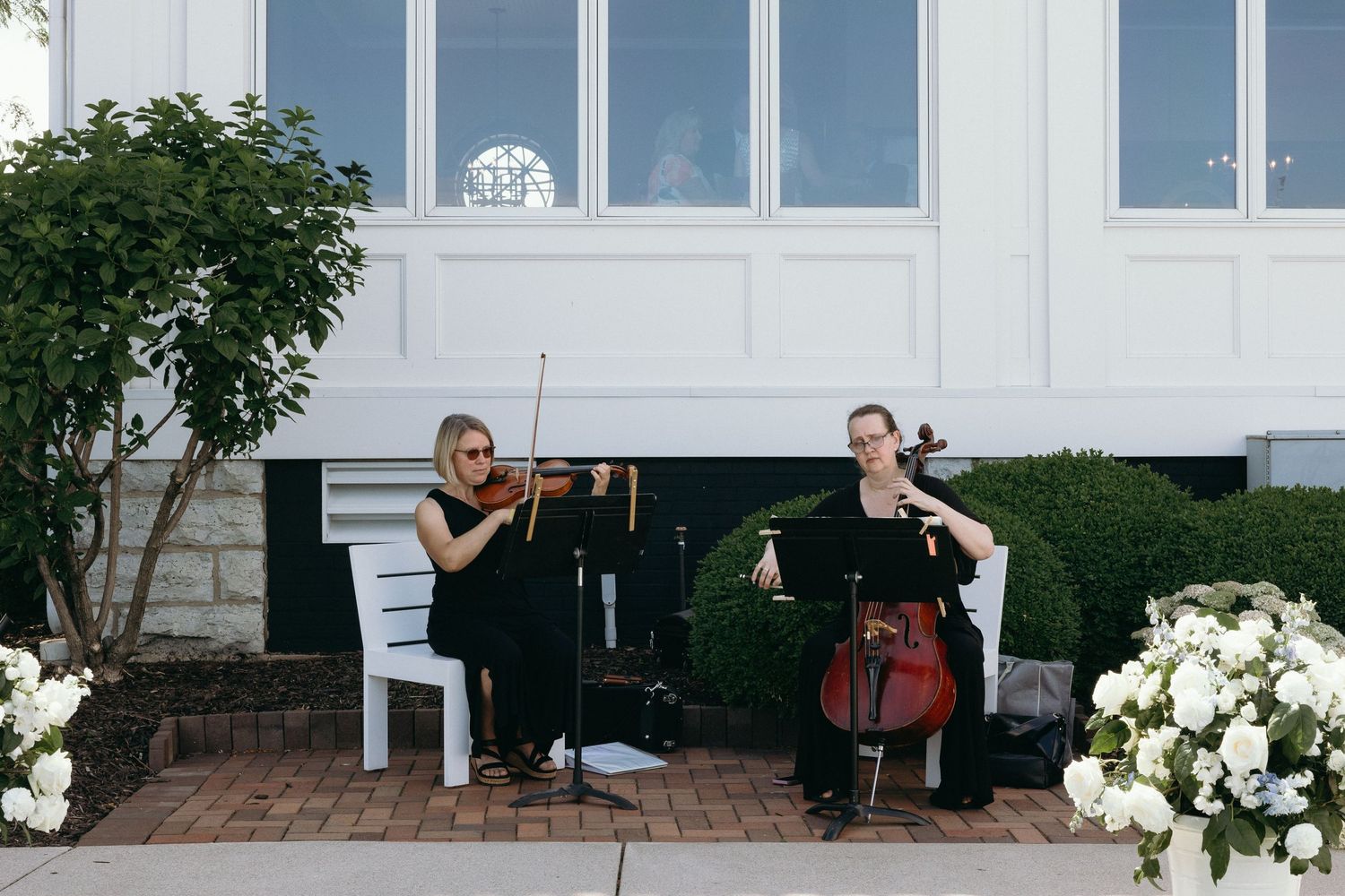 Musicians perform on violin and cello outside a white building with shrubs and white flowers in the garden.