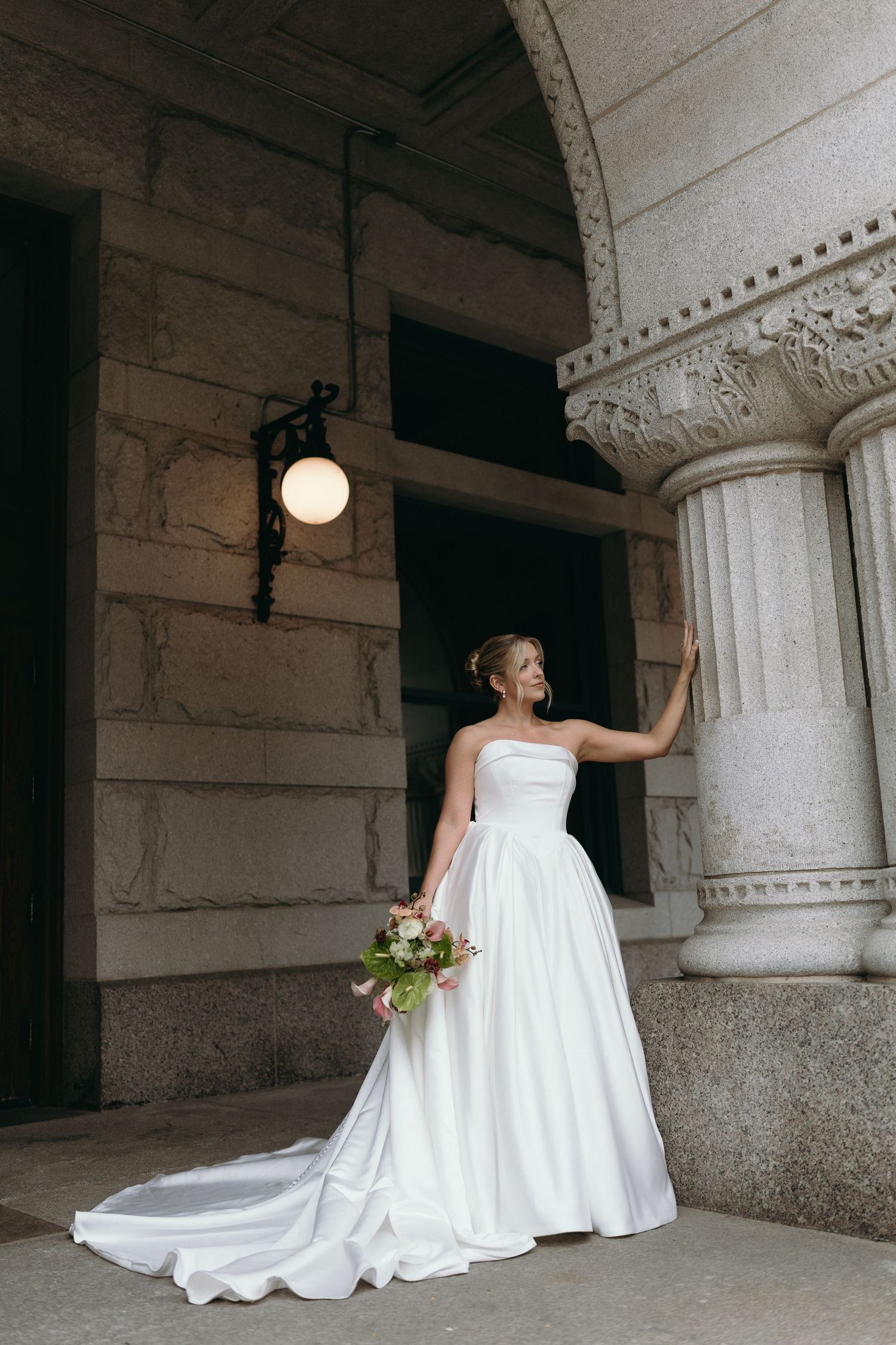 A bride in a flowing white wedding gown poses gracefully against ornate stone columns in a classical architectural setting.