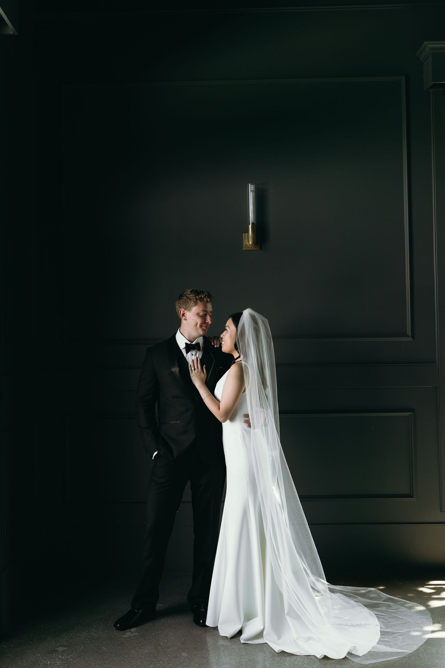 A bride in a flowing white gown and veil stands with a groom in black suit against a dark moody wall.