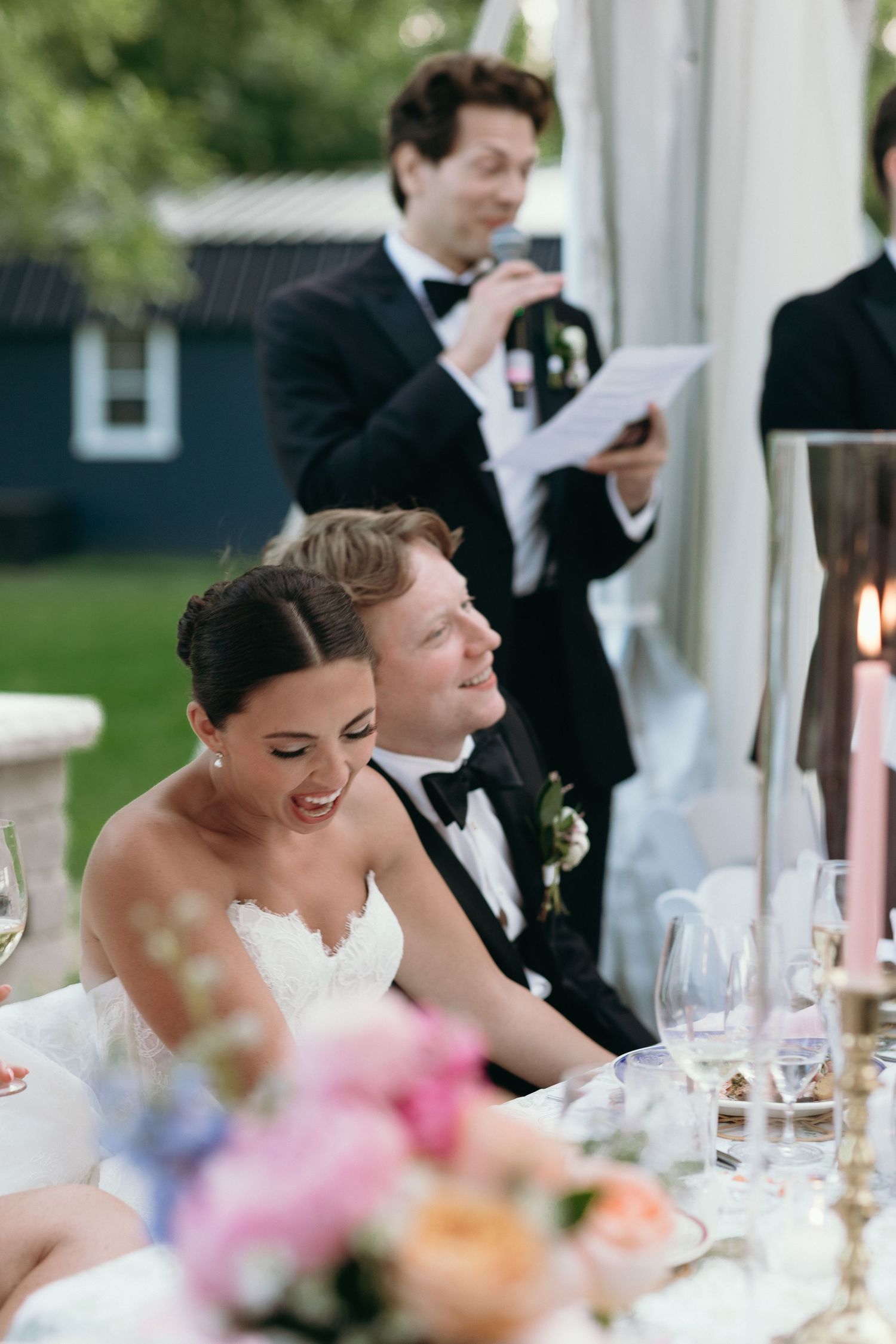 Smiling bride and groom listening to wedding speeches at elegantly decorated outdoor reception table.